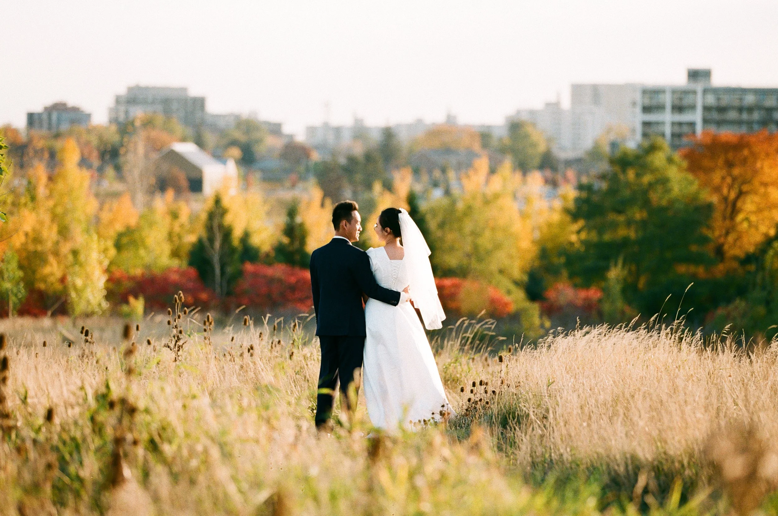 A bride and groom standing in a grassy field with autumn trees and city buildings in the background.