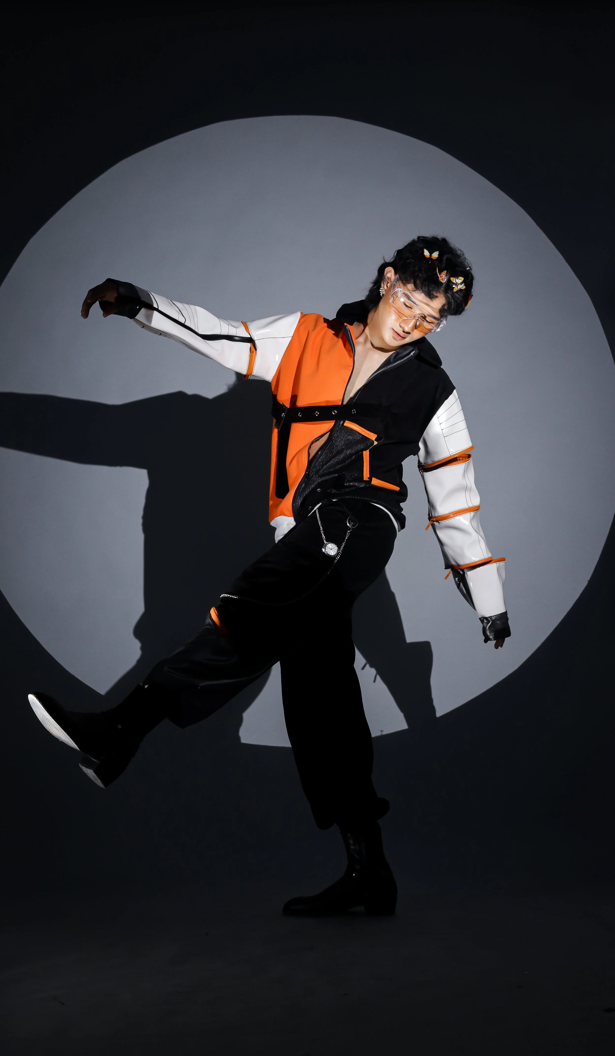 Person wearing orange and white jacket, black pants, and black boots, posing with one leg raised and arms outstretched against a gray background with a circular spotlight, wearing clear glasses and floral head accessories.