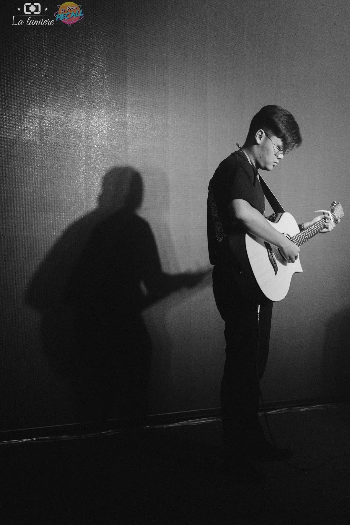 A young man playing an acoustic guitar against a textured wall, with his shadow cast on the wall.