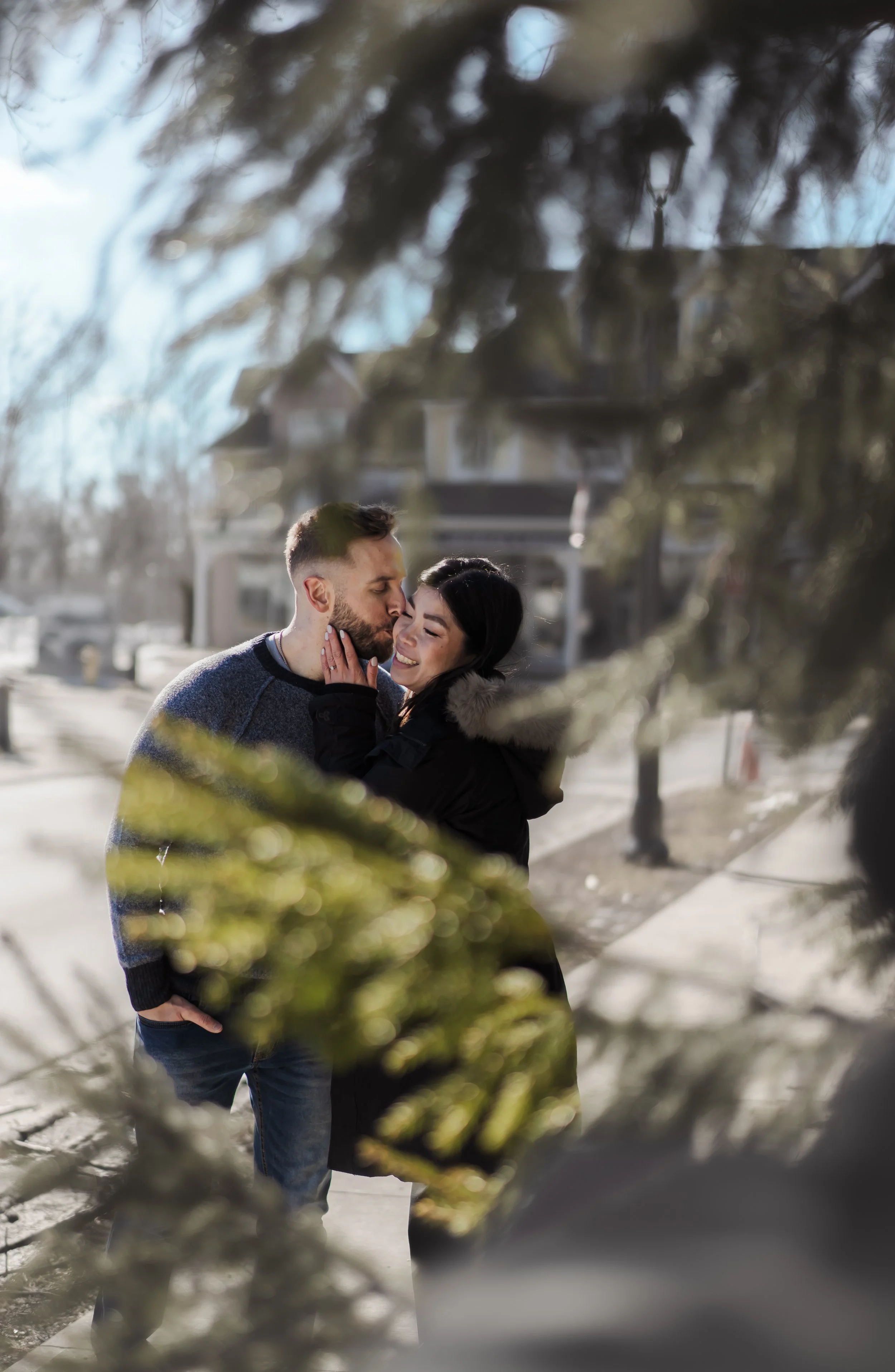 A young couple embracing outdoors in winter, smiling and touching foreheads, seen through the branches of a tree.