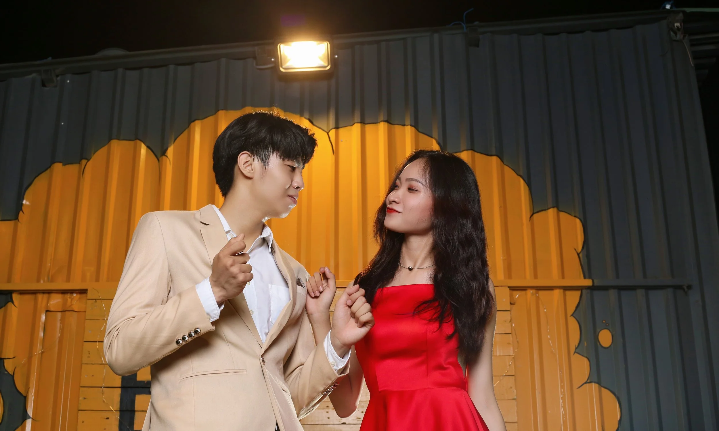A young man and woman stand close together in front of a colorful wall, smiling and looking into each other's eyes. The man is wearing a beige suit and the woman is wearing a red dress.
