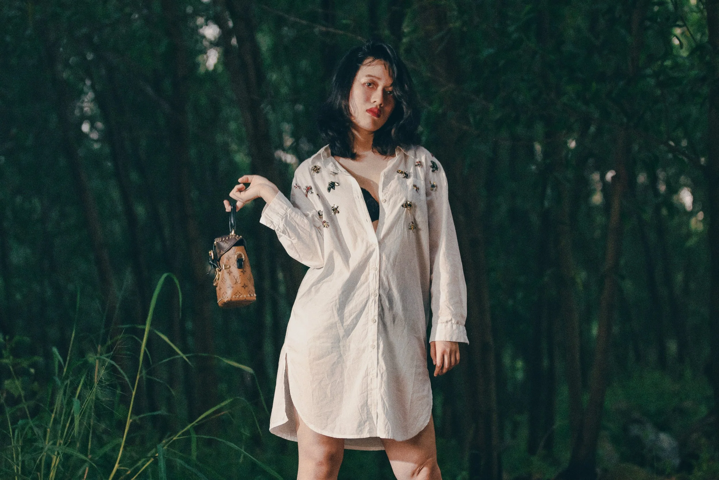 A woman with black hair wearing a white long shirt with embroidery, standing in a green, wooded outdoor area, holding a small bag.
