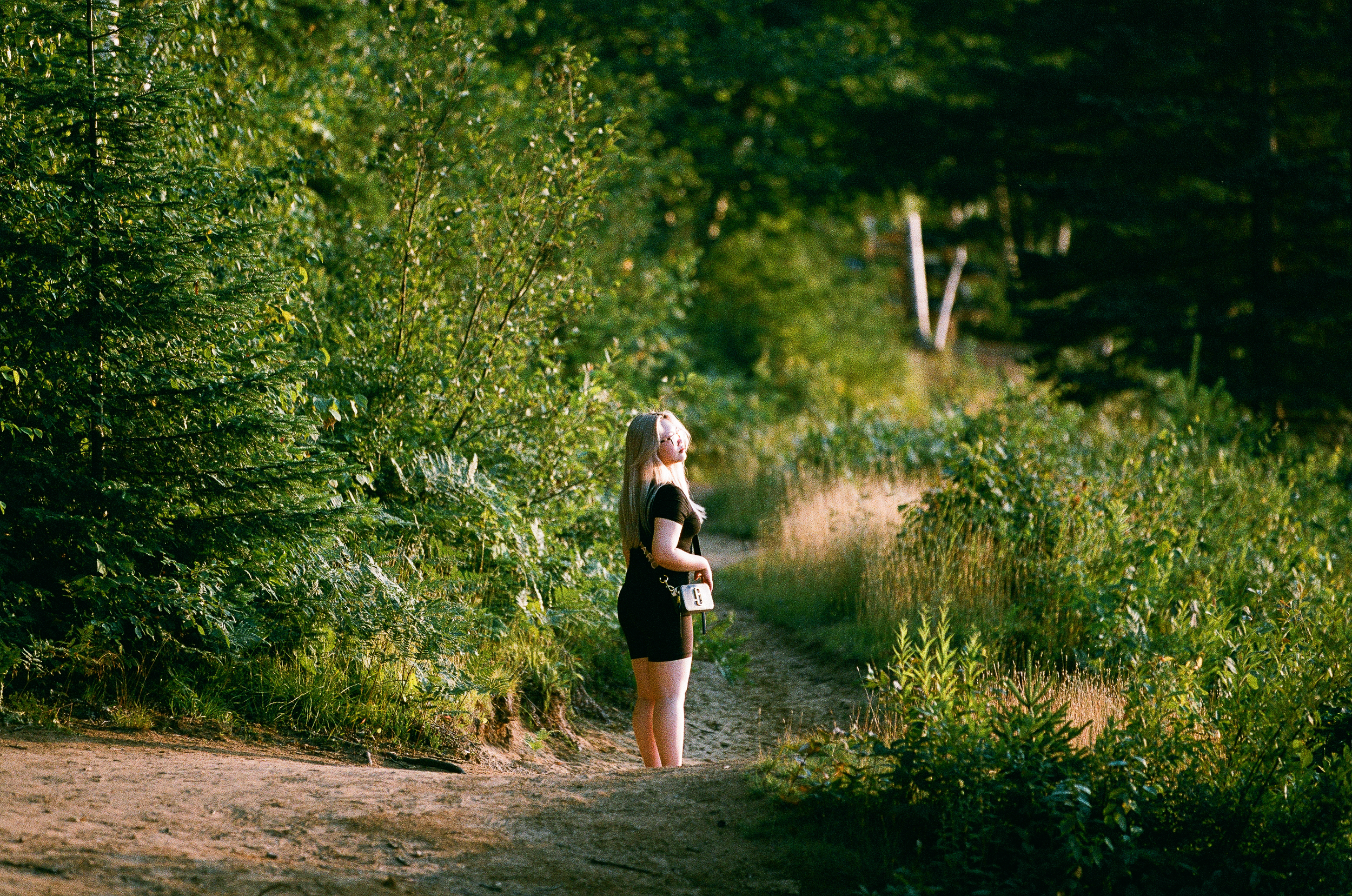 A woman in a black dress standing on a dirt trail in a lush green forest, with her eyes closed and face tilted up towards the sunlight.