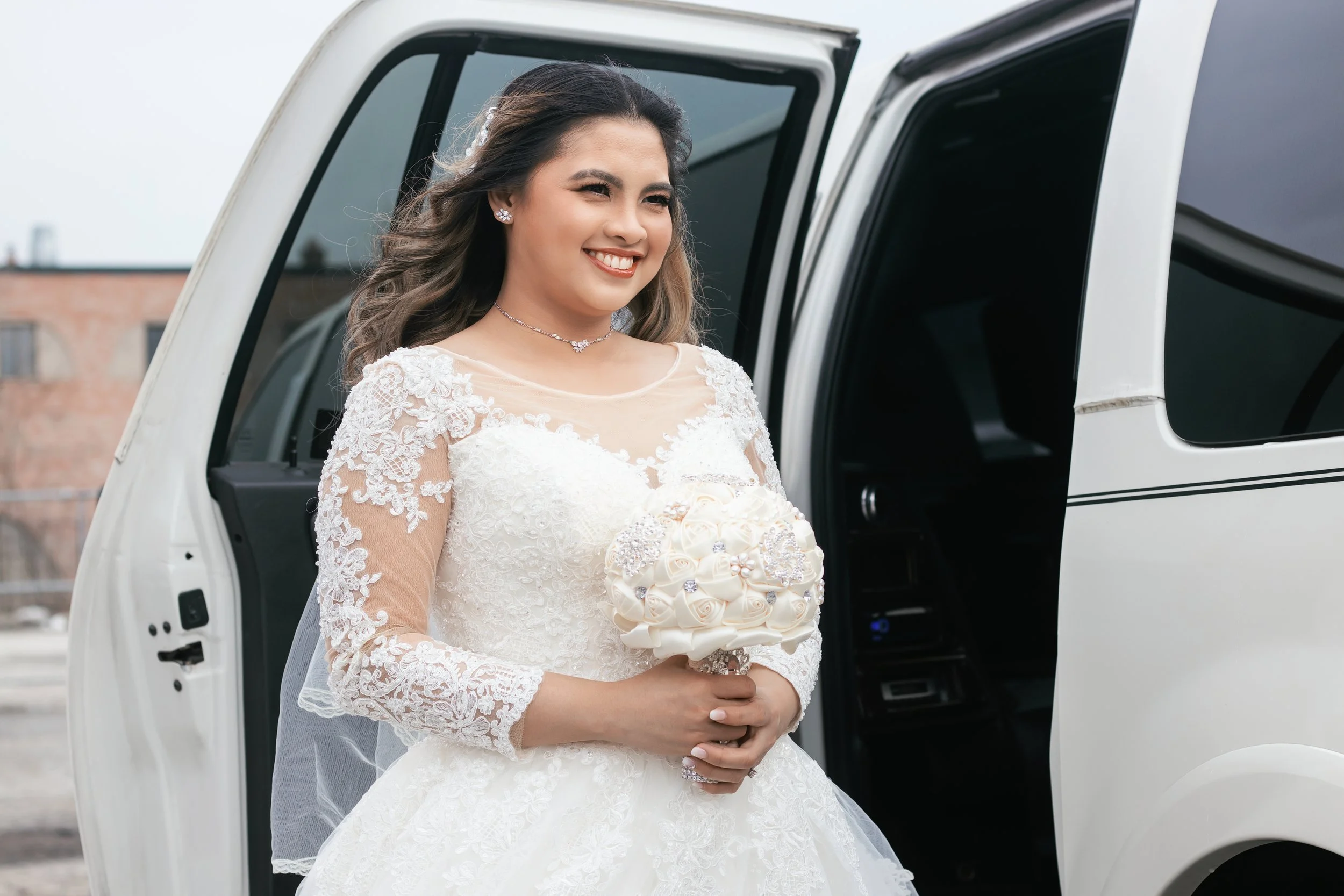 A bride dressed in a white lace wedding gown holding a bouquet of white roses, standing beside a white limo.