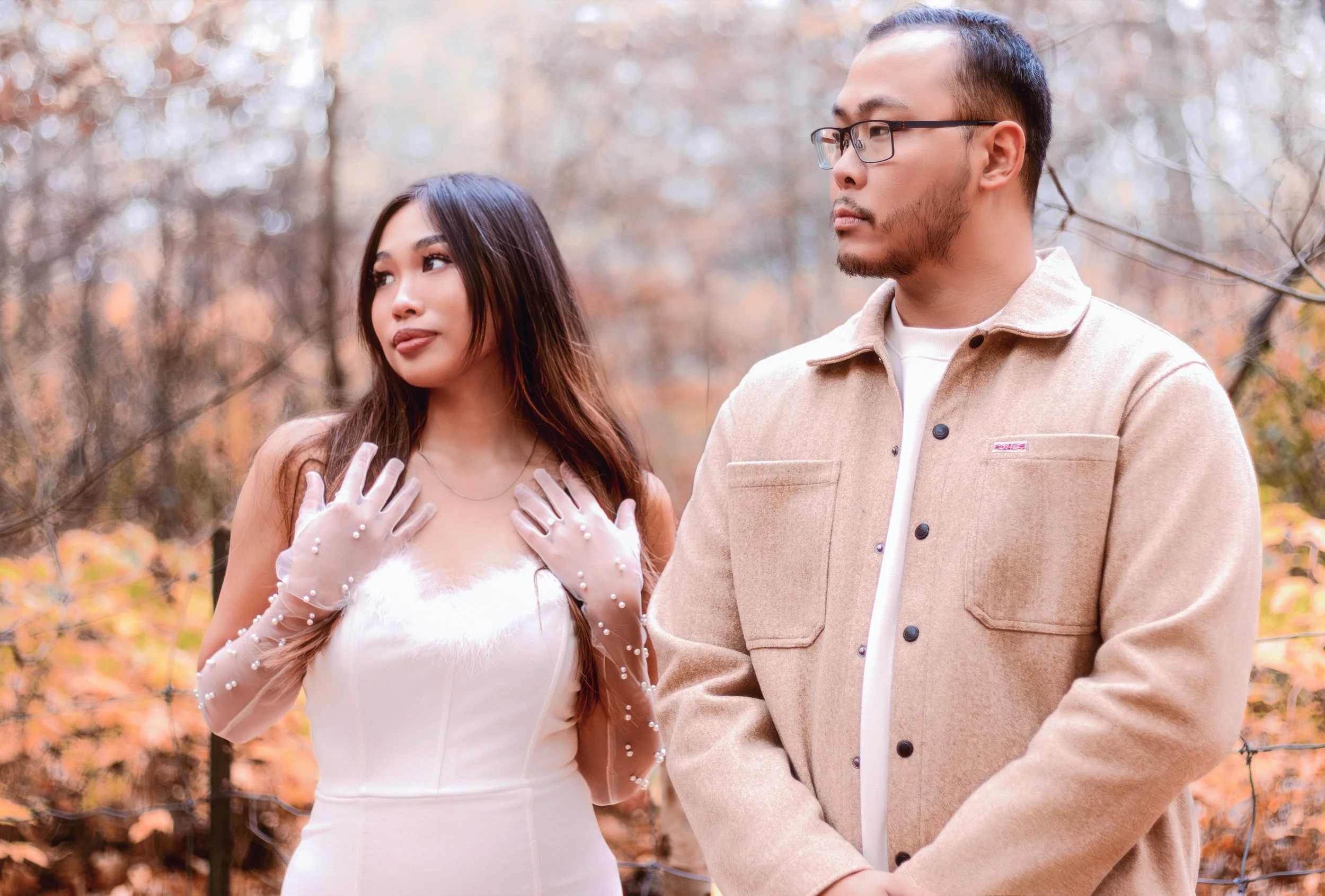 A woman in a white dress with pearl-embellished sheer gloves standing outdoors in autumn, and a man in a beige coat with glasses standing beside her in a forested area.