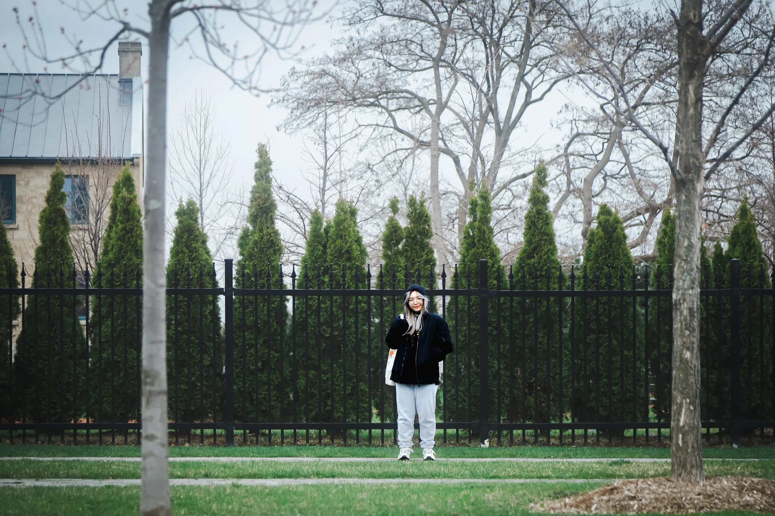 A woman with blonde hair wearing a black coat and gray sweatpants standing in front of a black metal fence with tall green evergreen trees behind her. She is holding a phone to her ear and smiling, with bare trees and a building with a chimney in the
