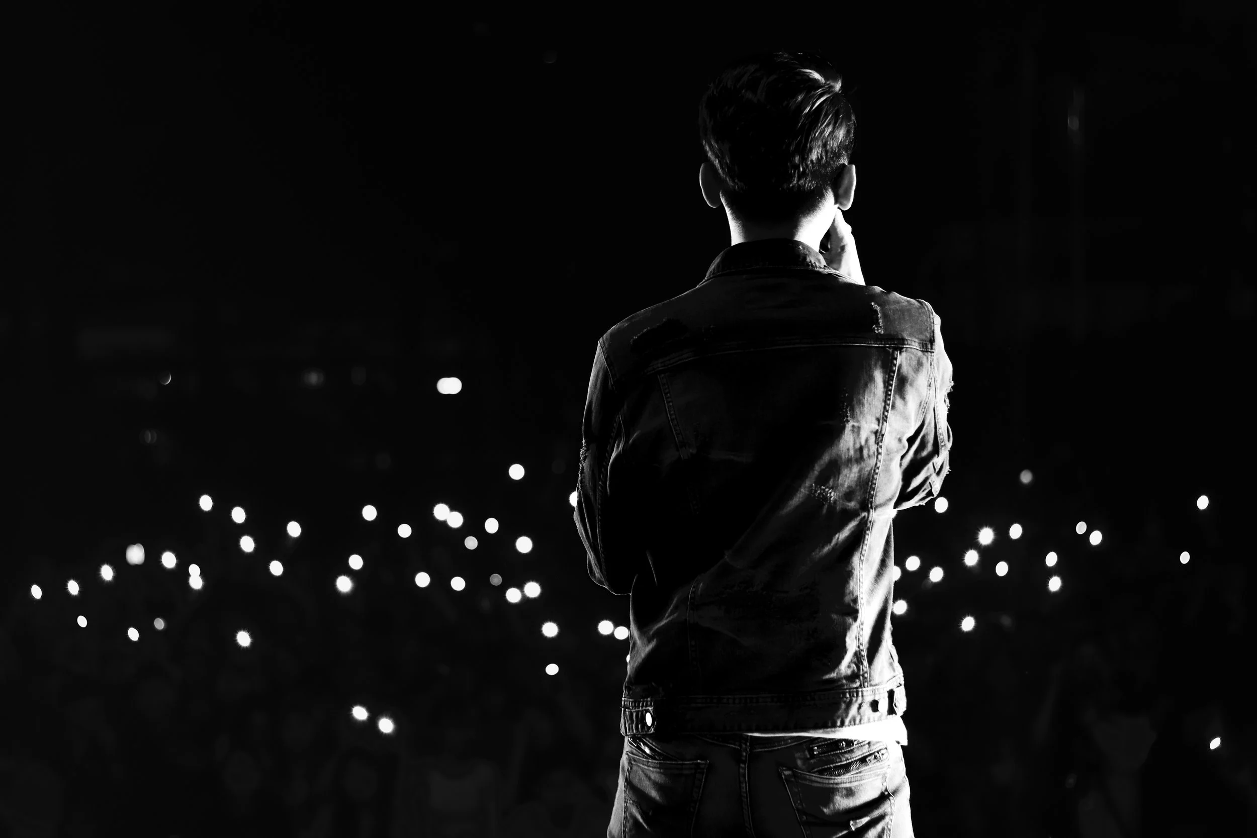 A man with short hair wearing a leather jacket stands with his back to the camera, looking at a dark night sky illuminated by small bright lights or stars.