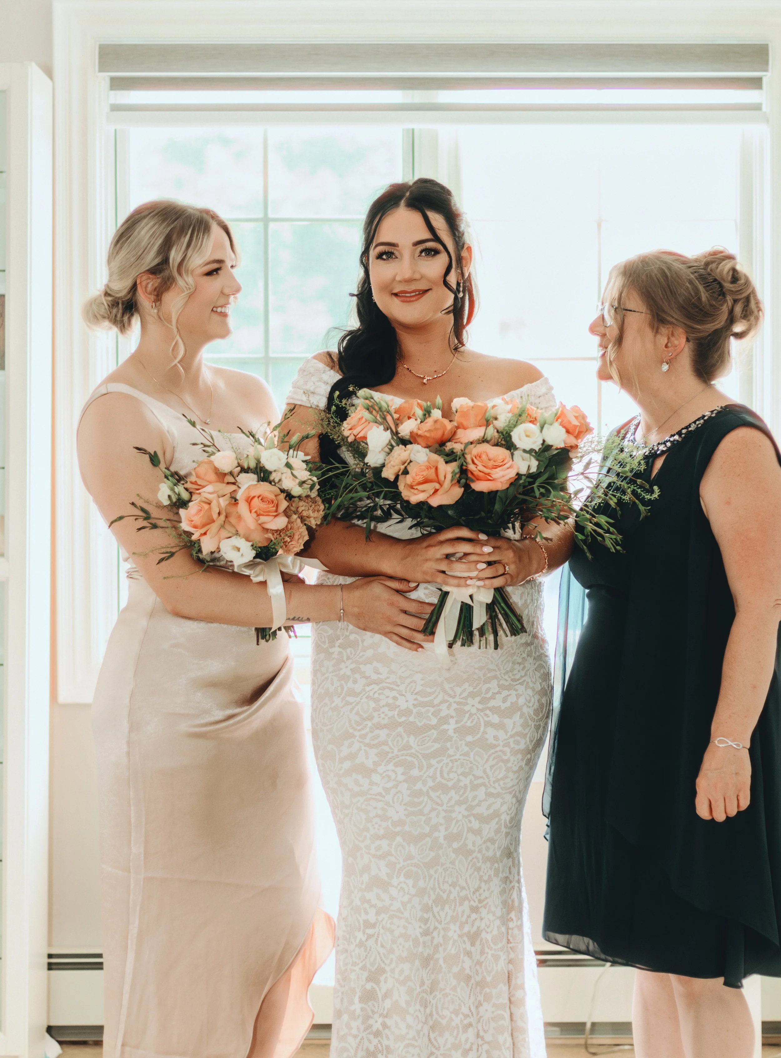 A bride in a white lace wedding dress holding a bouquet of peach and white flowers, standing between her bridesmaids and mother, also holding bouquets, in front of a sunlit window during a wedding celebration.