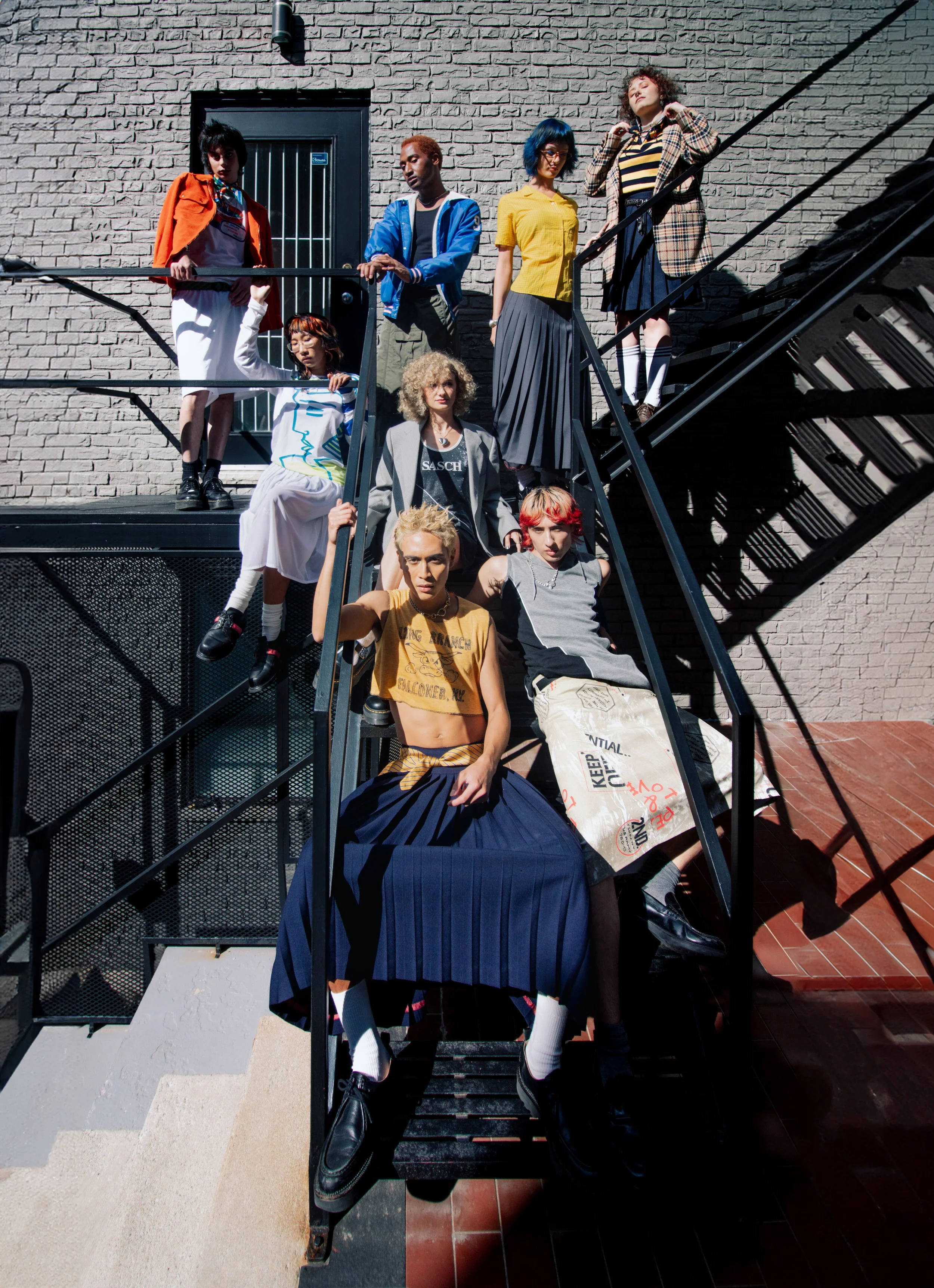 A group of nine diverse young people posing on a metal staircase outdoors against a brick wall. They are dressed in colorful, trendy clothing and exhibit various poses and expressions.