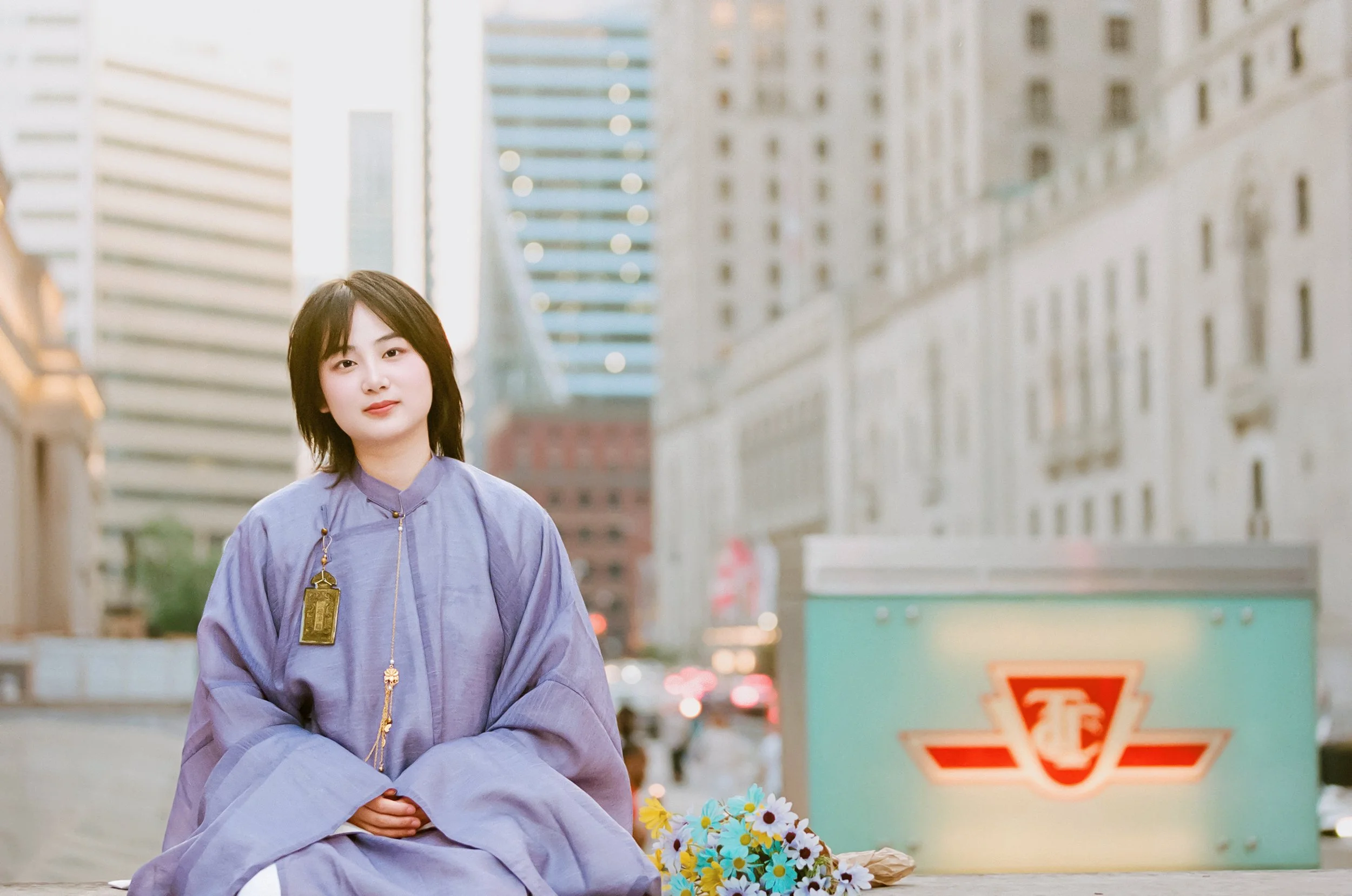 A young woman with short dark hair and a gentle smile sitting outdoors in a city setting during daylight. She is wearing a loose, lavender-colored tunic with long sleeves and a gold pendant. There are flowers placed beside her on the ground. Tall bui