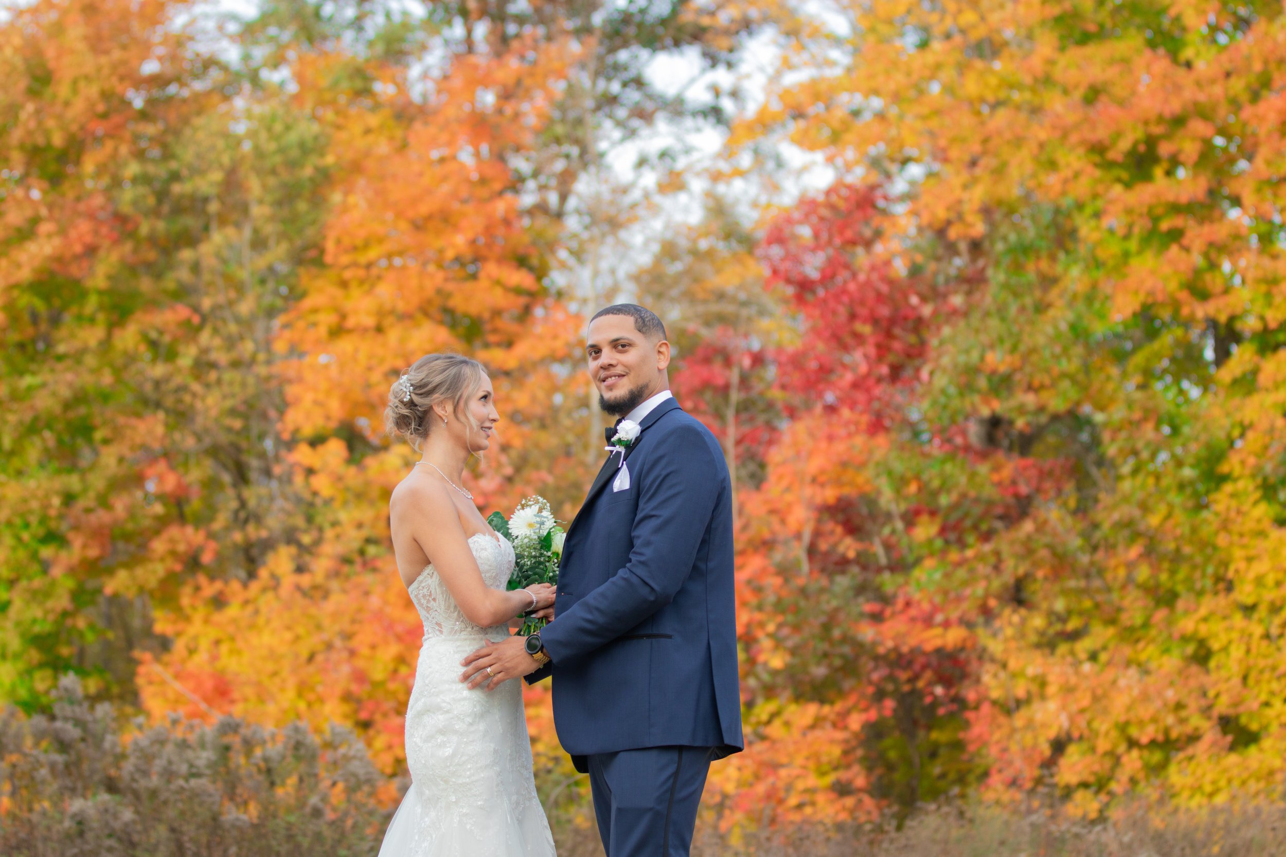 A bride and groom standing outdoors during a wedding, holding hands, with colorful autumn leaves in the background.