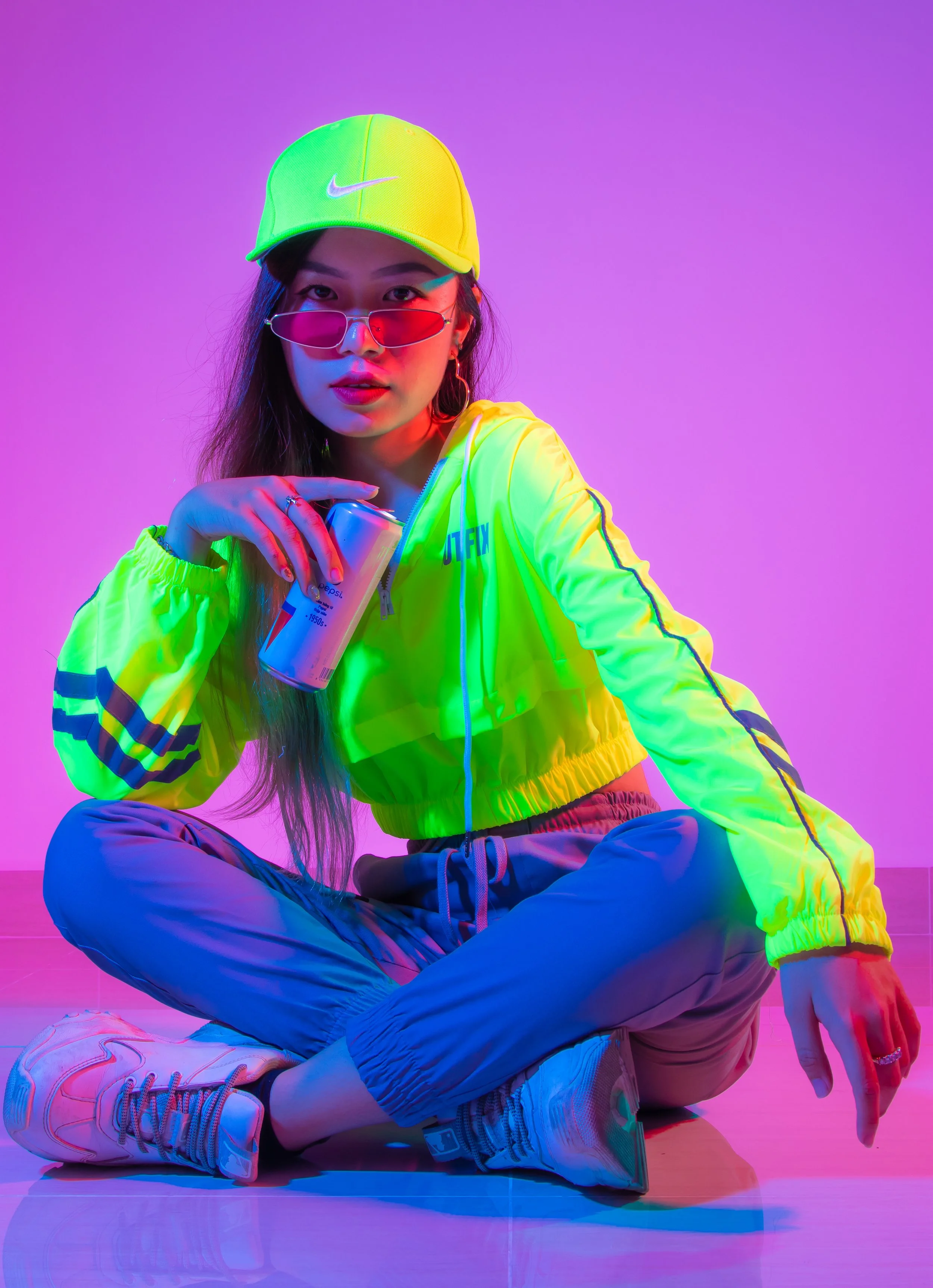 A woman sitting cross-legged on the floor holding a soda can, wearing a neon yellow Nike cap, neon yellow jacket, gray pants, and white sneakers, with pink sunglasses, against a colorful pink and purple gradient background.