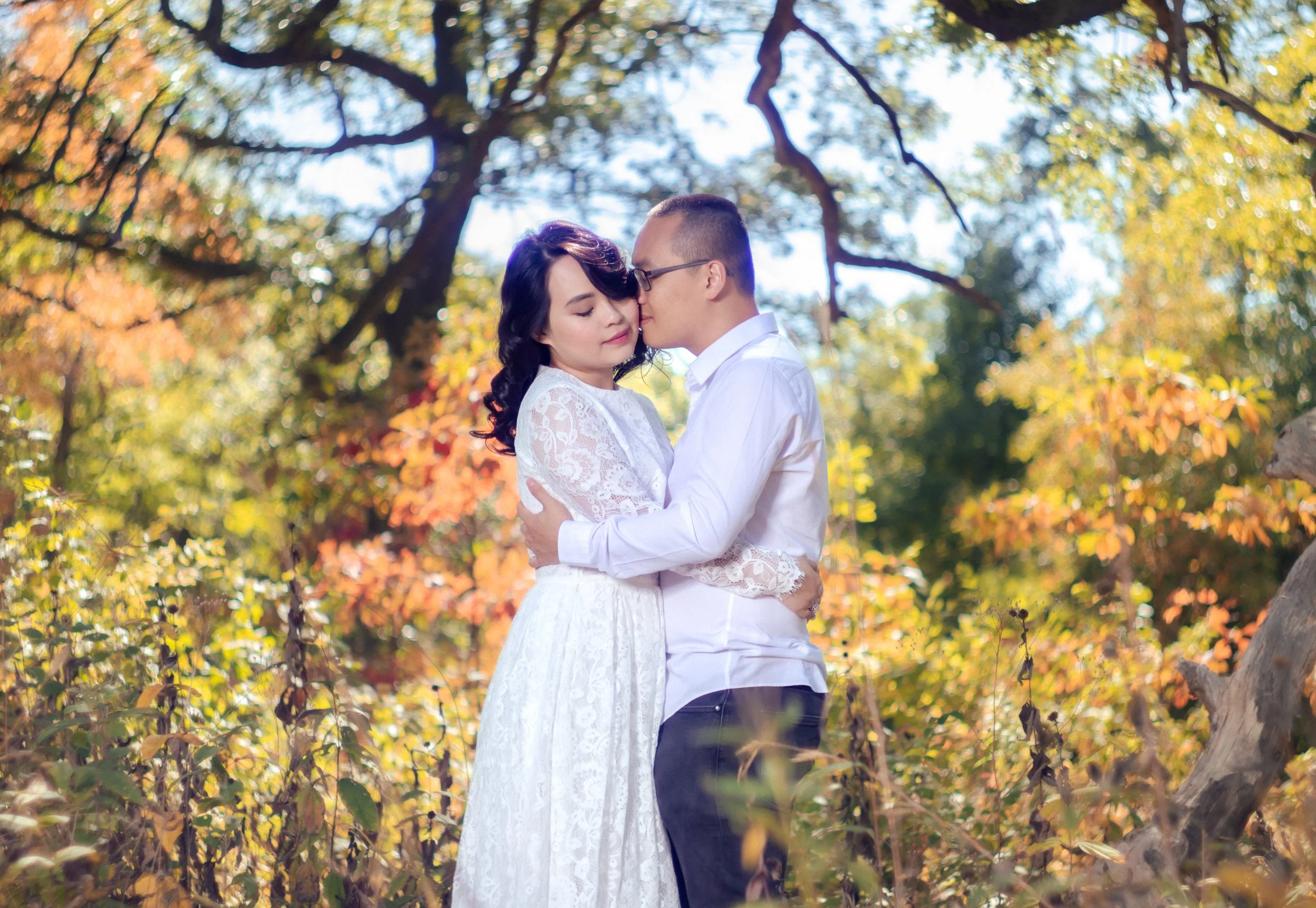 A young couple dressed in white, embracing closely outdoors among fall foliage, with trees and colorful leaves in the background.