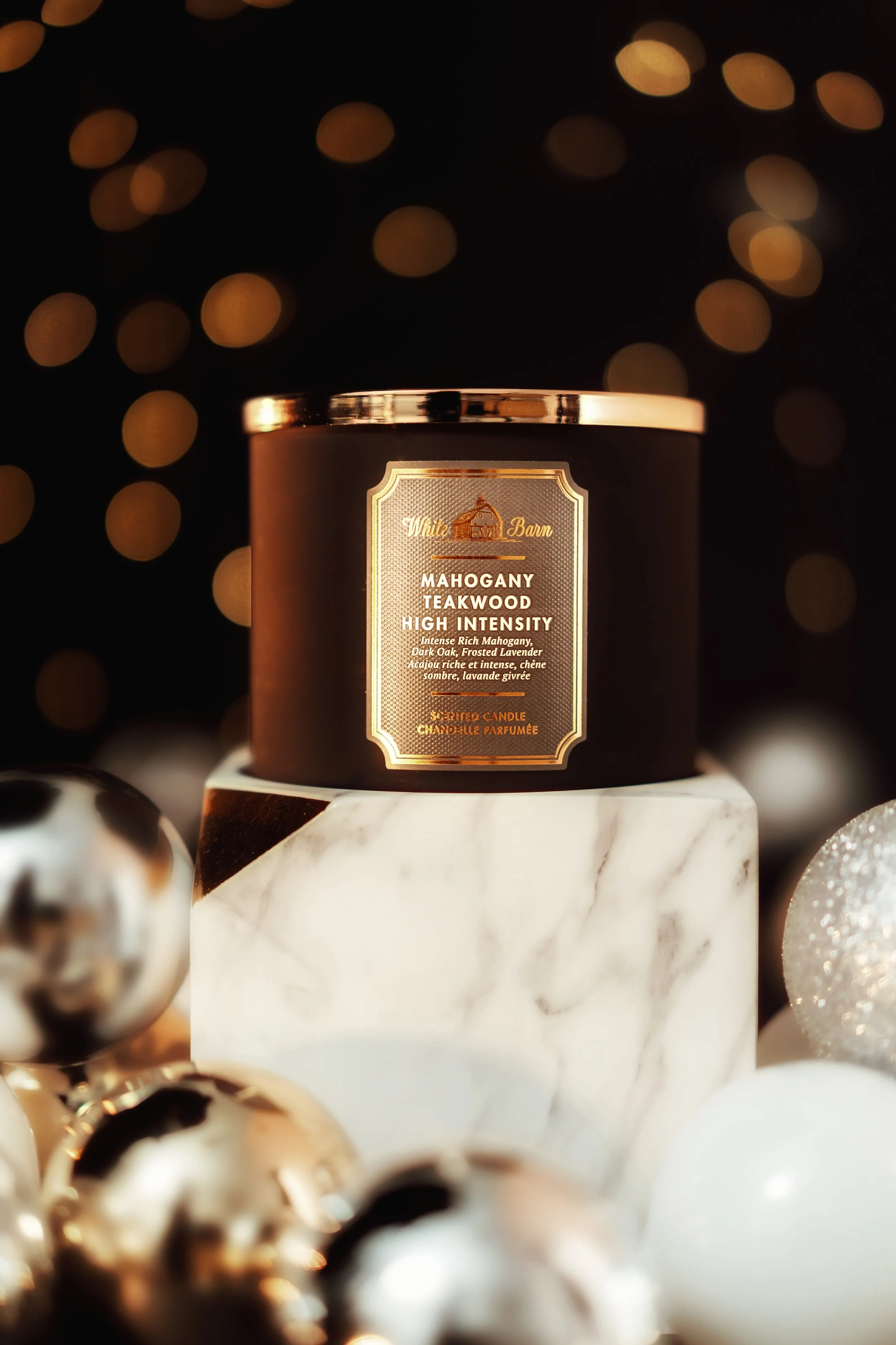 A black jar of White Barn mahogany teakwood high intensity scented candle on a marble stand, surrounded by gold and white decorative balls, with blurred festive lights in the background.