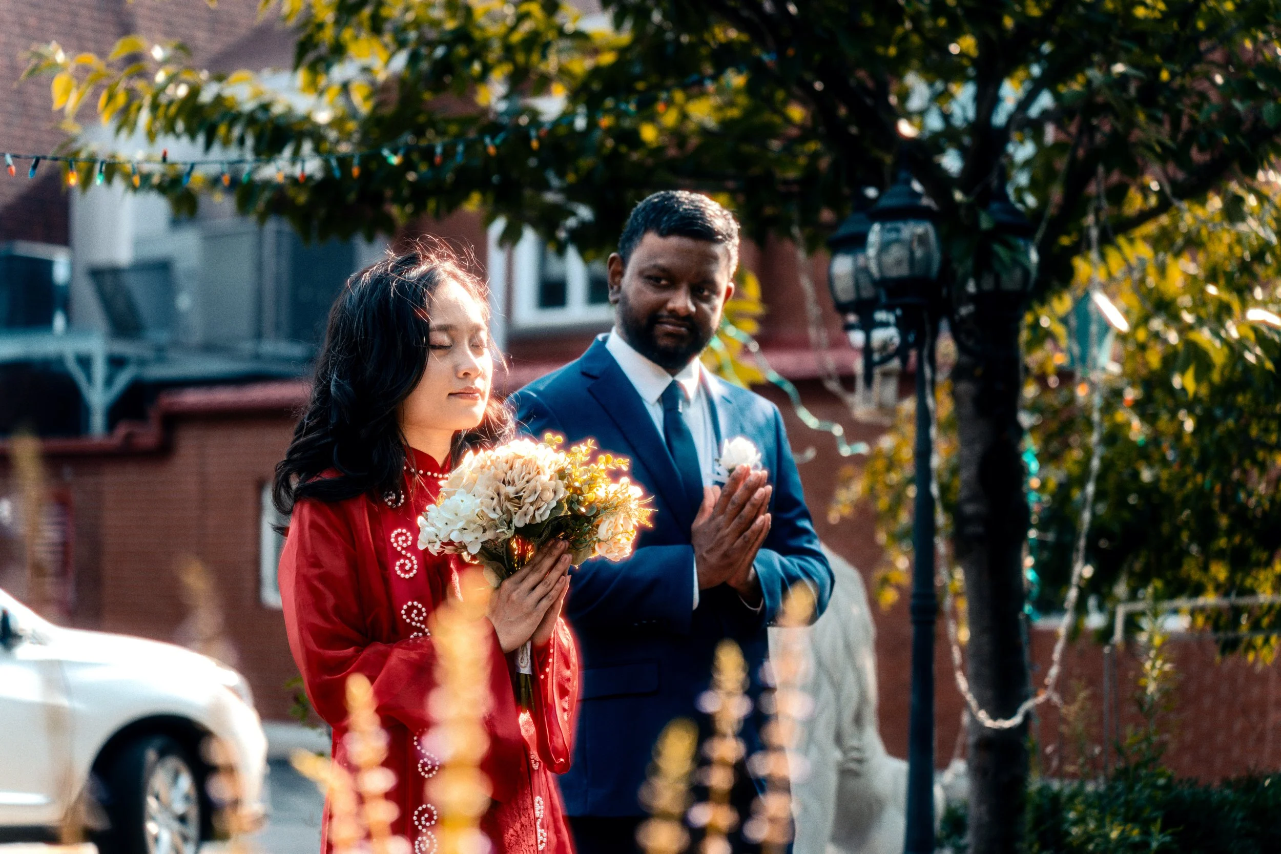 A woman with long dark hair and a man with short dark hair and a beard are standing outdoors during the daytime, with their hands pressed together in a prayer-like gesture. The woman is holding a bouquet of white and light-colored flowers, and the ma