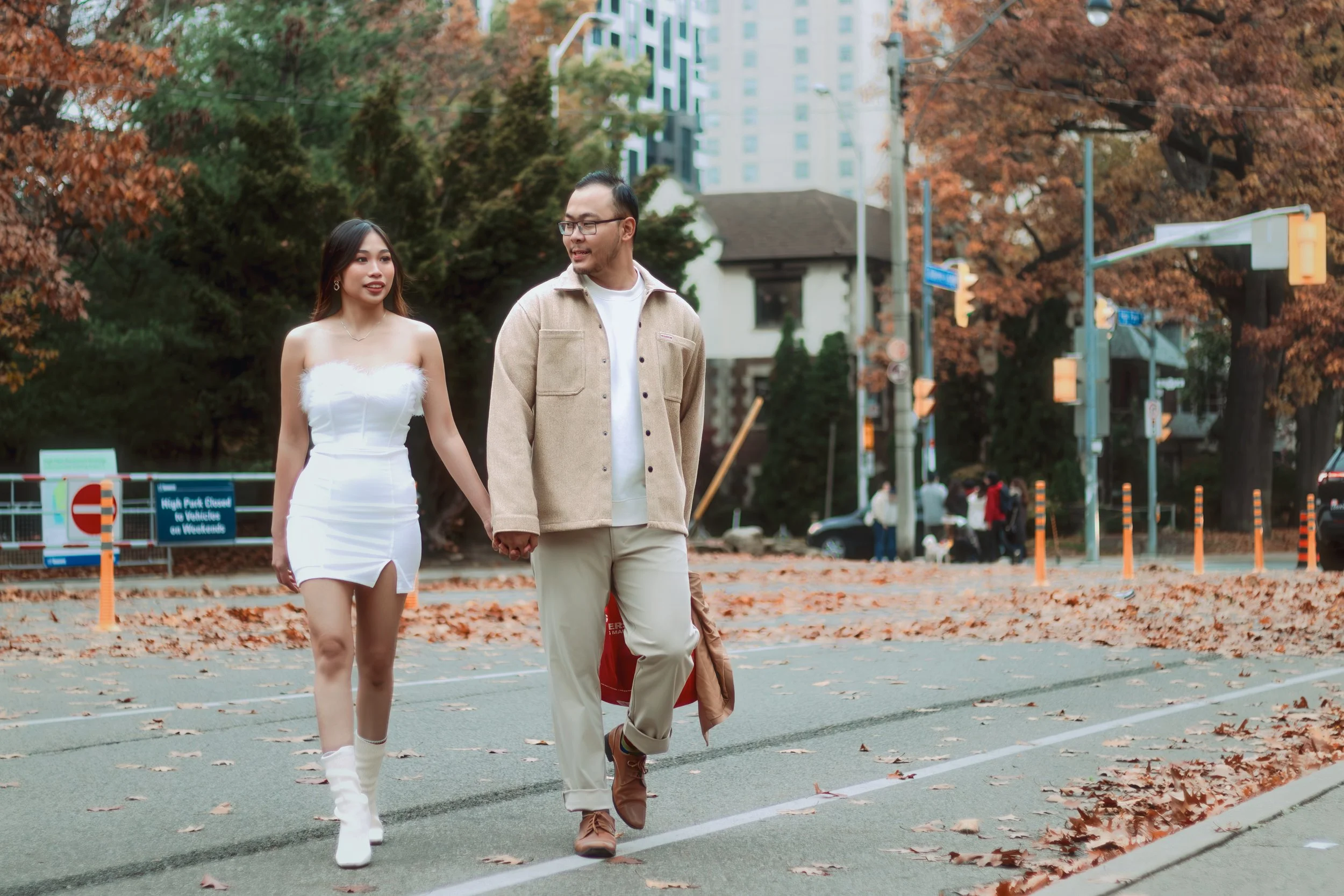 A young woman and a man are walking hand in hand outdoors on a city street with fallen autumn leaves, trees, and tall buildings in the background.
