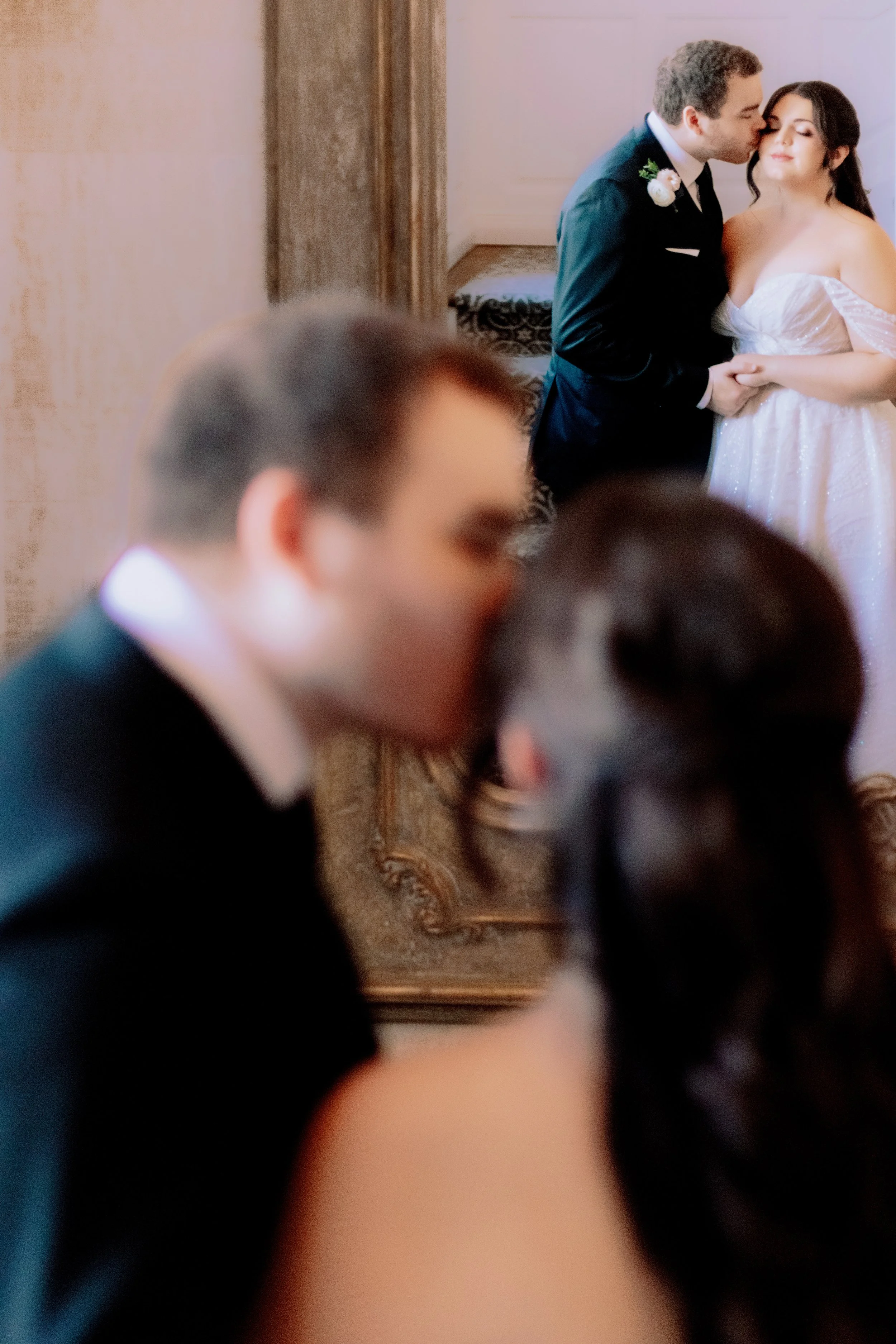 A bride and groom share a kiss in the background, while a couple in the foreground appears to be kissing during a wedding ceremony.