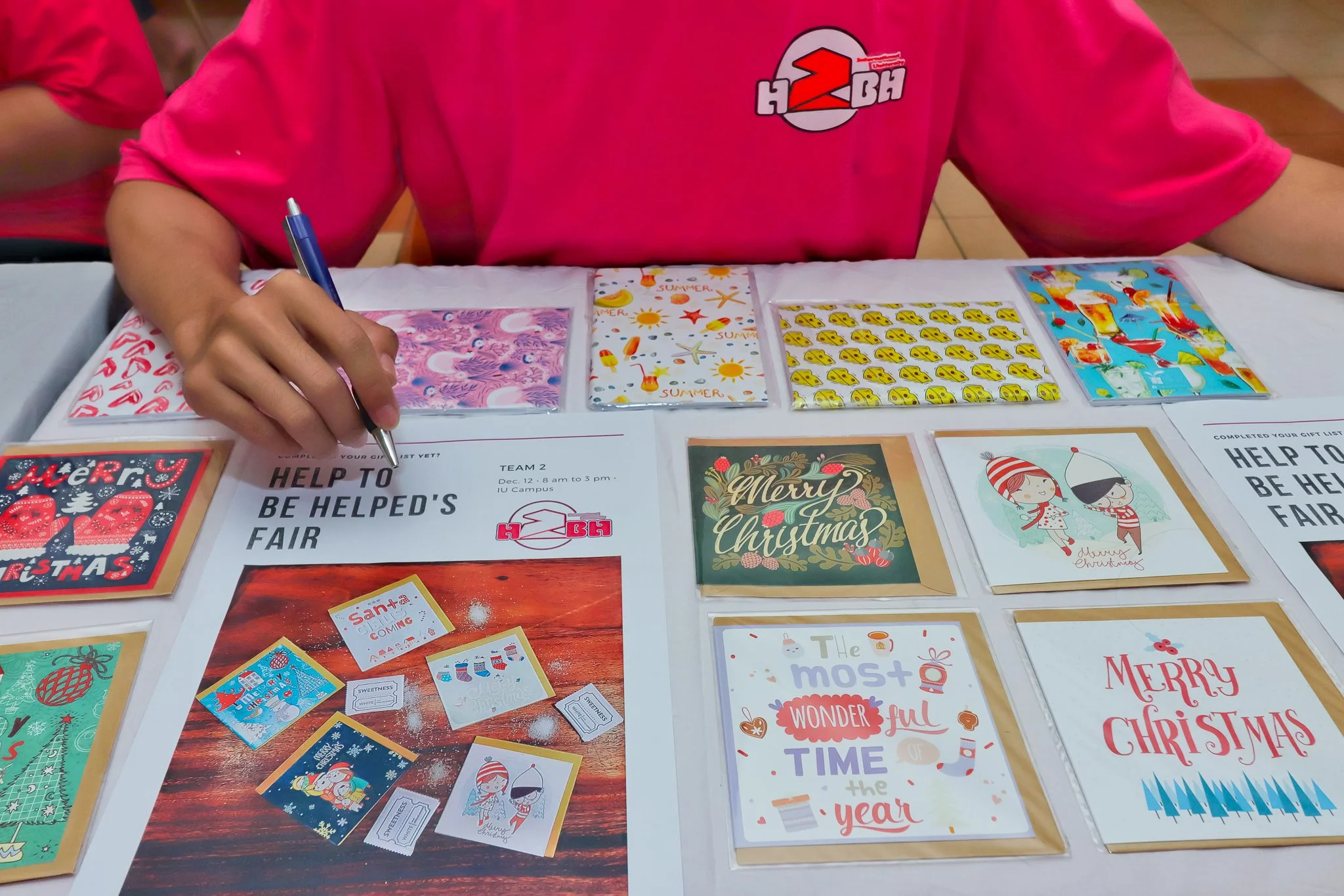 Person sitting at a table, writing with a pen, with Christmas cards and decorative paper on the table.