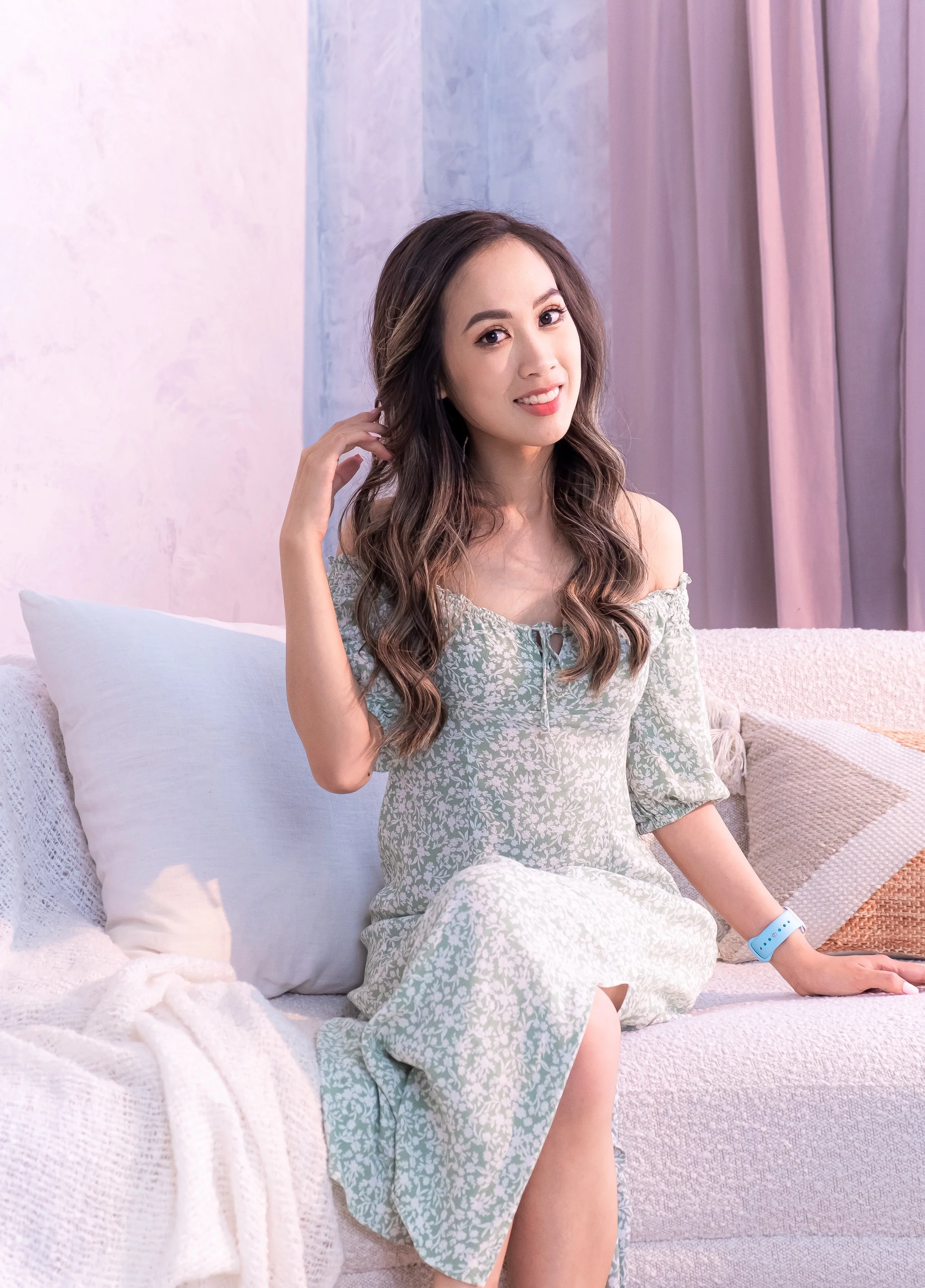A young woman with long wavy hair sitting on a white couch in a softly lit room with pastel-colored walls and pink curtains, smiling and looking at the camera.