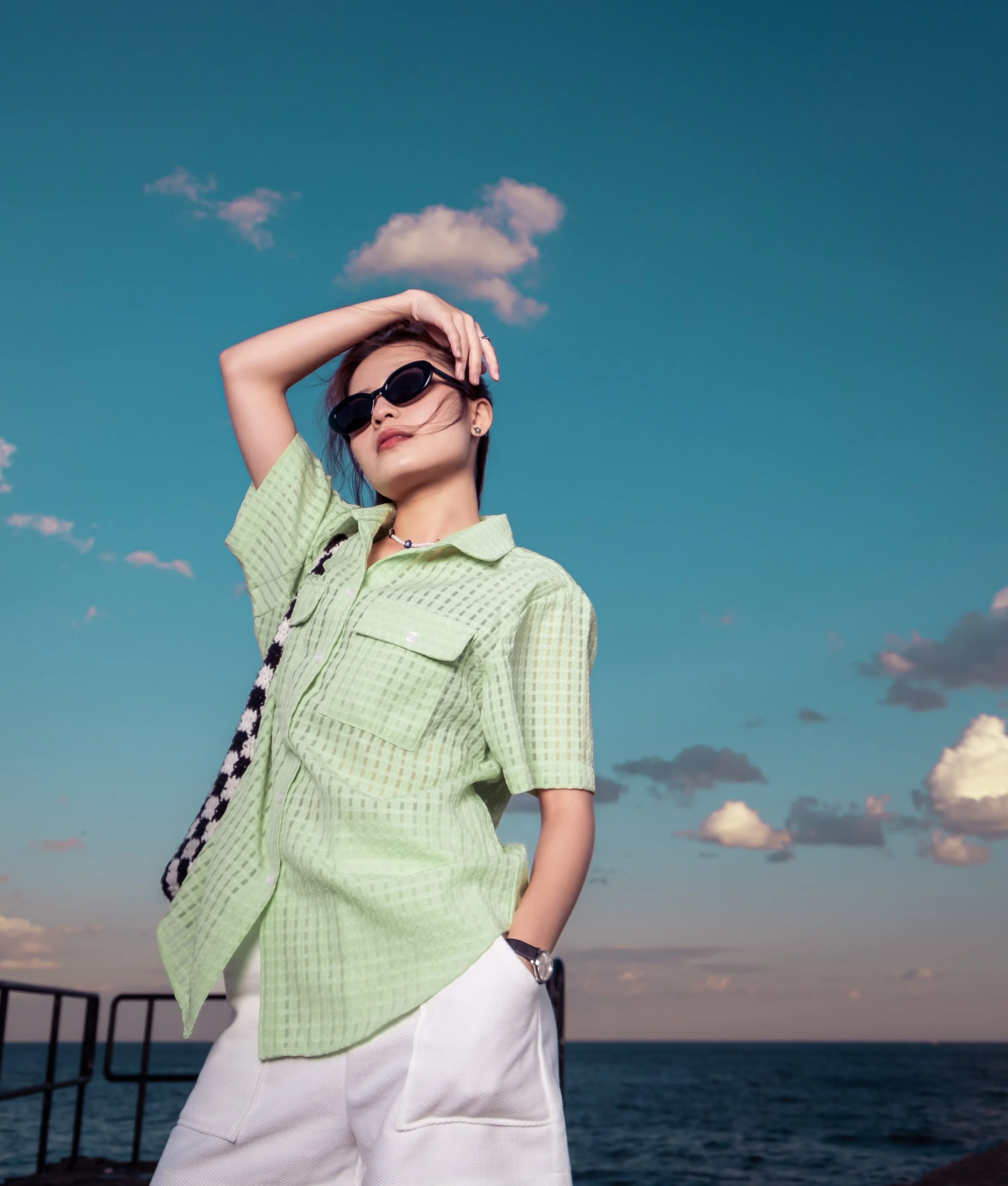 Woman wearing sunglasses, green checkered shirt, and white shorts standing outdoors by the water under a partly cloudy sky