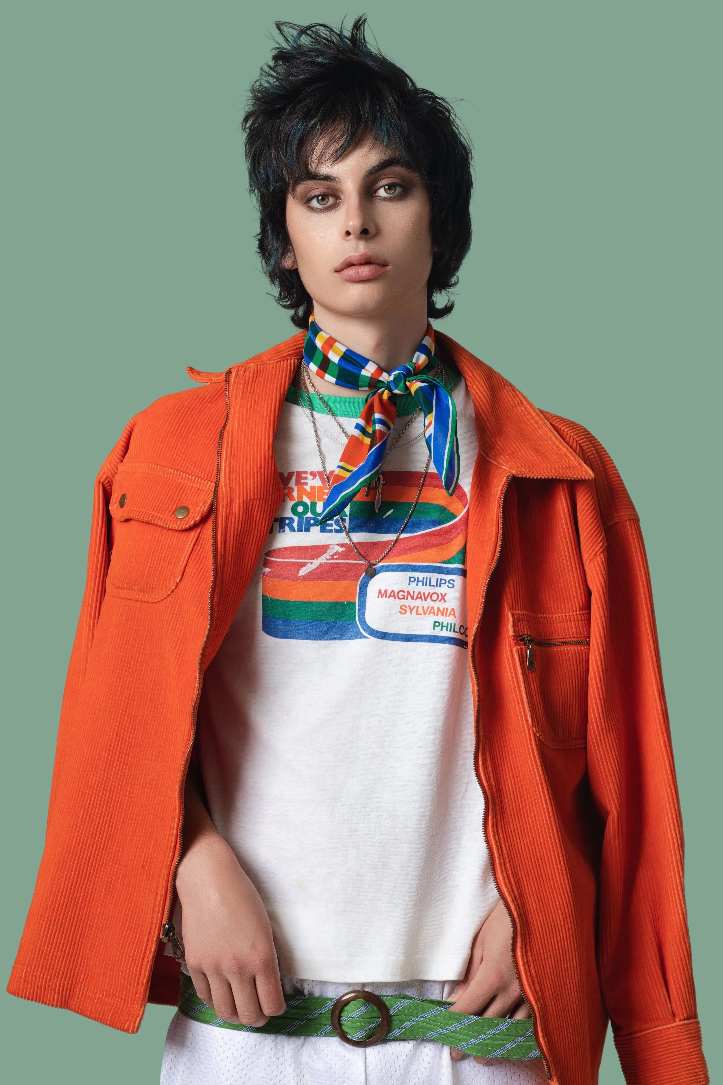 A young person with short, tousled black hair wearing a vibrant orange jacket, a white graphic t-shirt, green and white striped pants, and a multicolored neck scarf, standing against a muted green background.