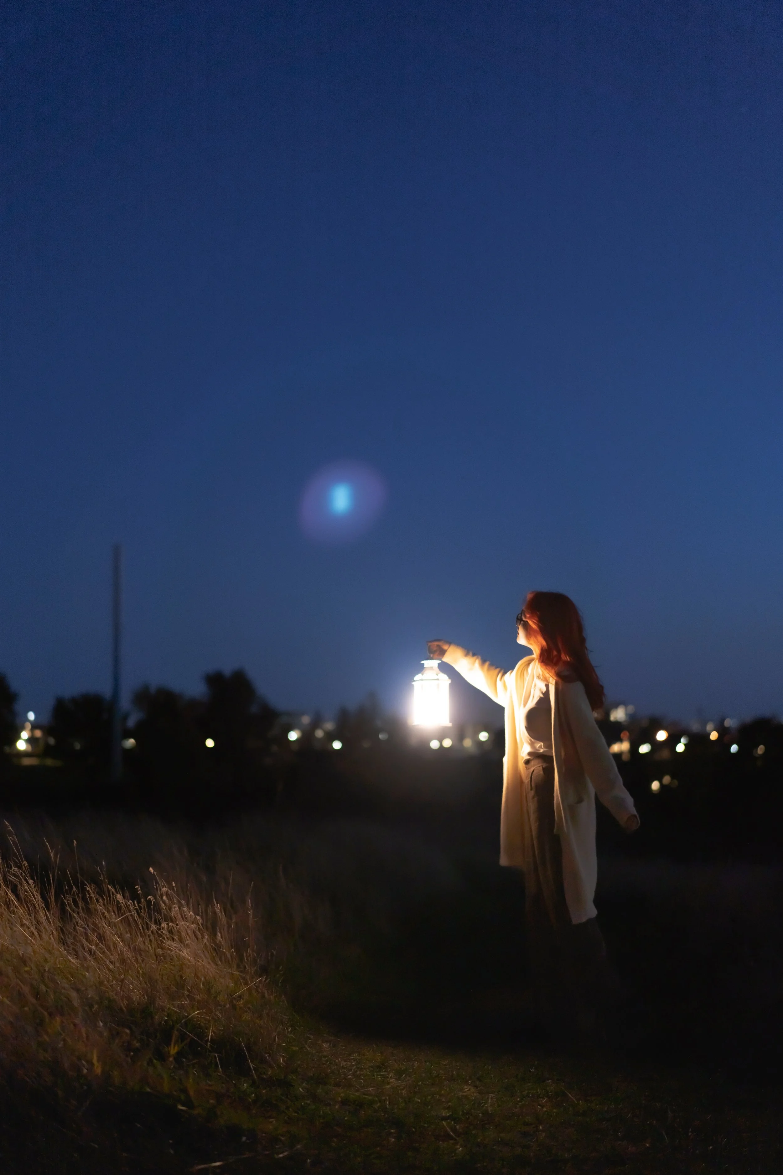 A woman in a coat standing outdoors at night, holding a glowing lantern, with a bright blue star or planet in the sky above.