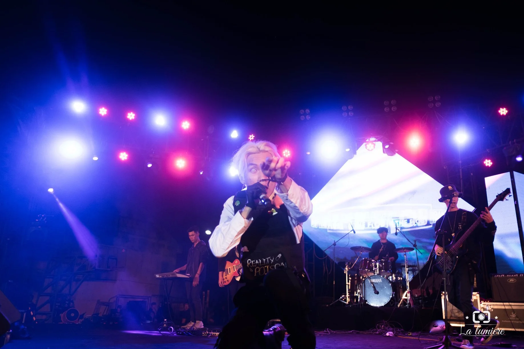 A band performing on stage with a lead singer in front, pointing towards the camera, under colorful stage lights with a large LED screen in the background.