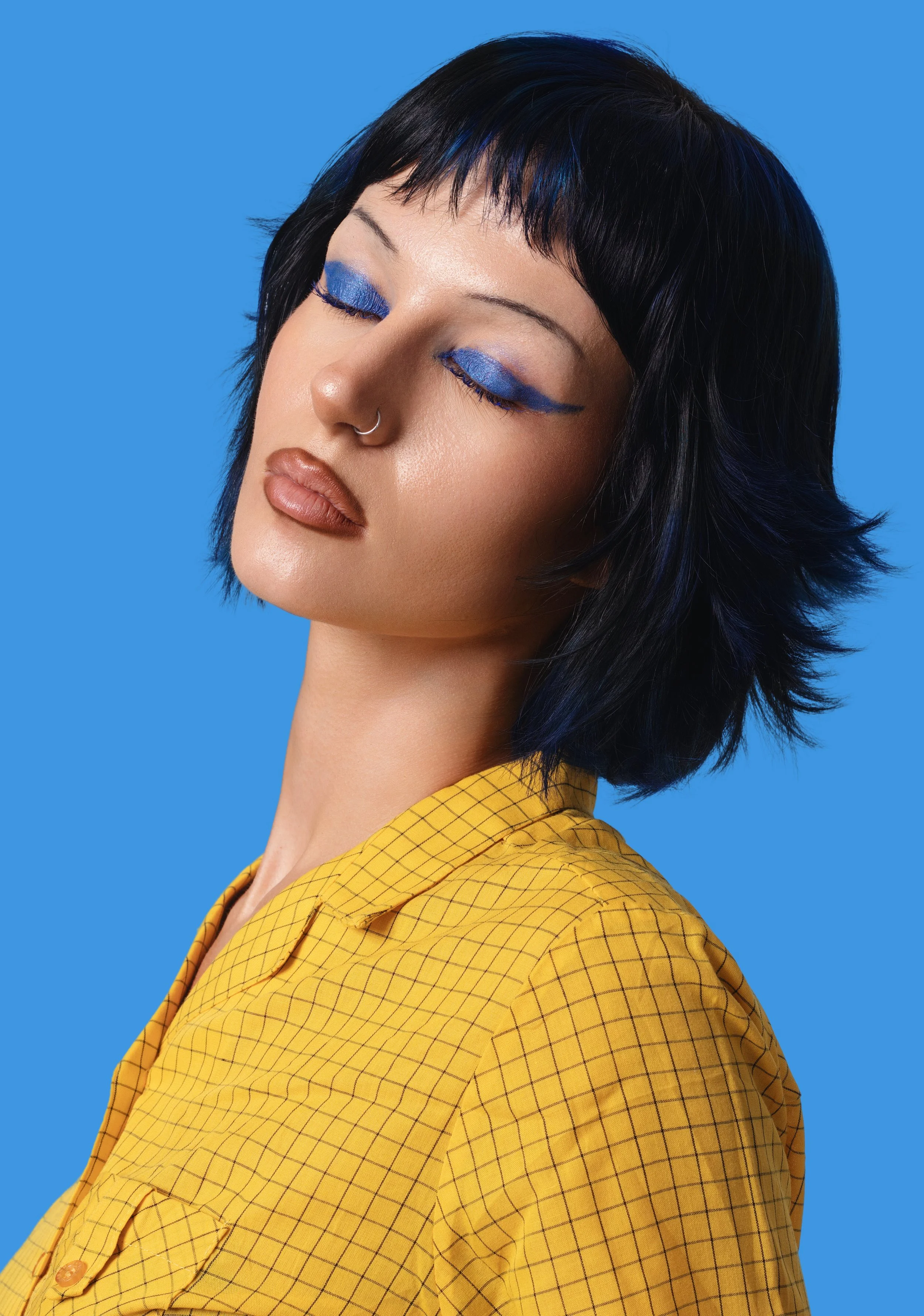 A woman with short black hair with blue highlights, wearing blue eyeshadow with winged eyeliner, nude lipstick, and a yellow checkered shirt against a blue background.