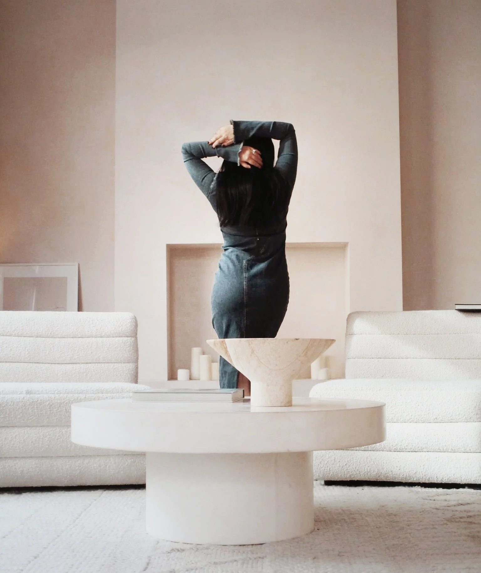 A woman with dark hair standing in a modern living room with her back to the camera, arms raised and hands behind her head, dressed in a gray dress with leather sleeves.