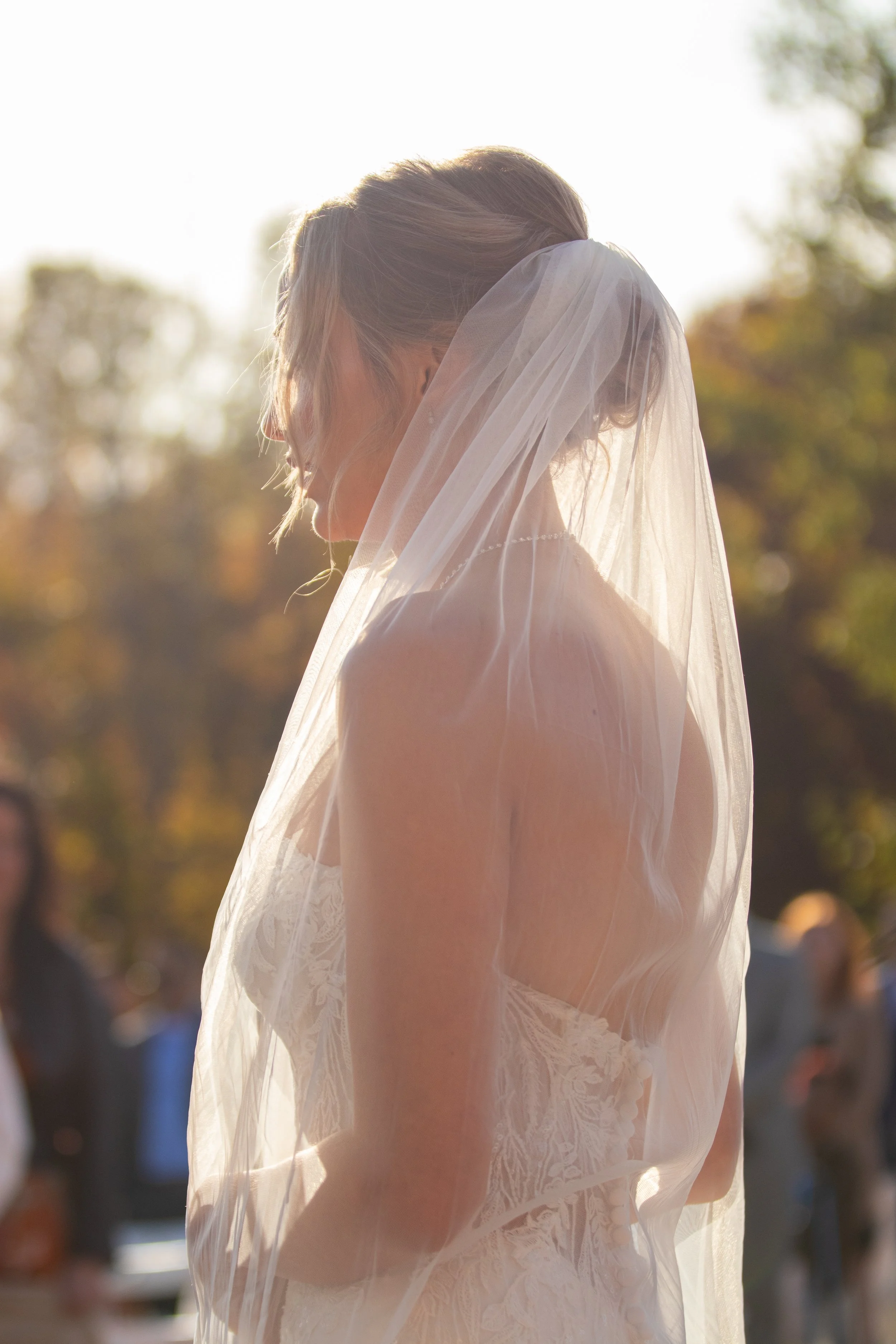 A bride wearing a wedding dress and veil, standing outdoors with sunlight behind her, with blurred people and trees in the background.