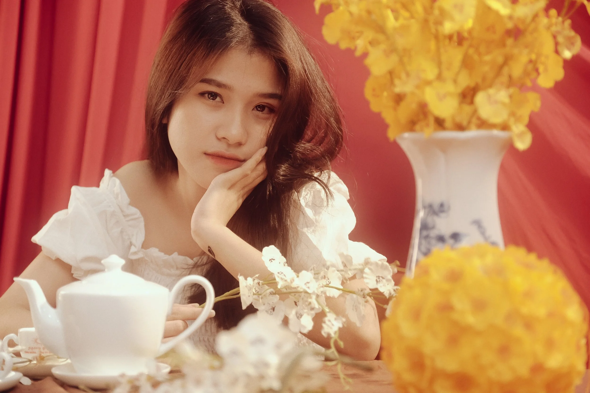 A woman with wavy brown hair and fair skin resting her face on her hand at a tea table with white teapot, white flowers, and yellow flowers in a vase against a red curtain background.