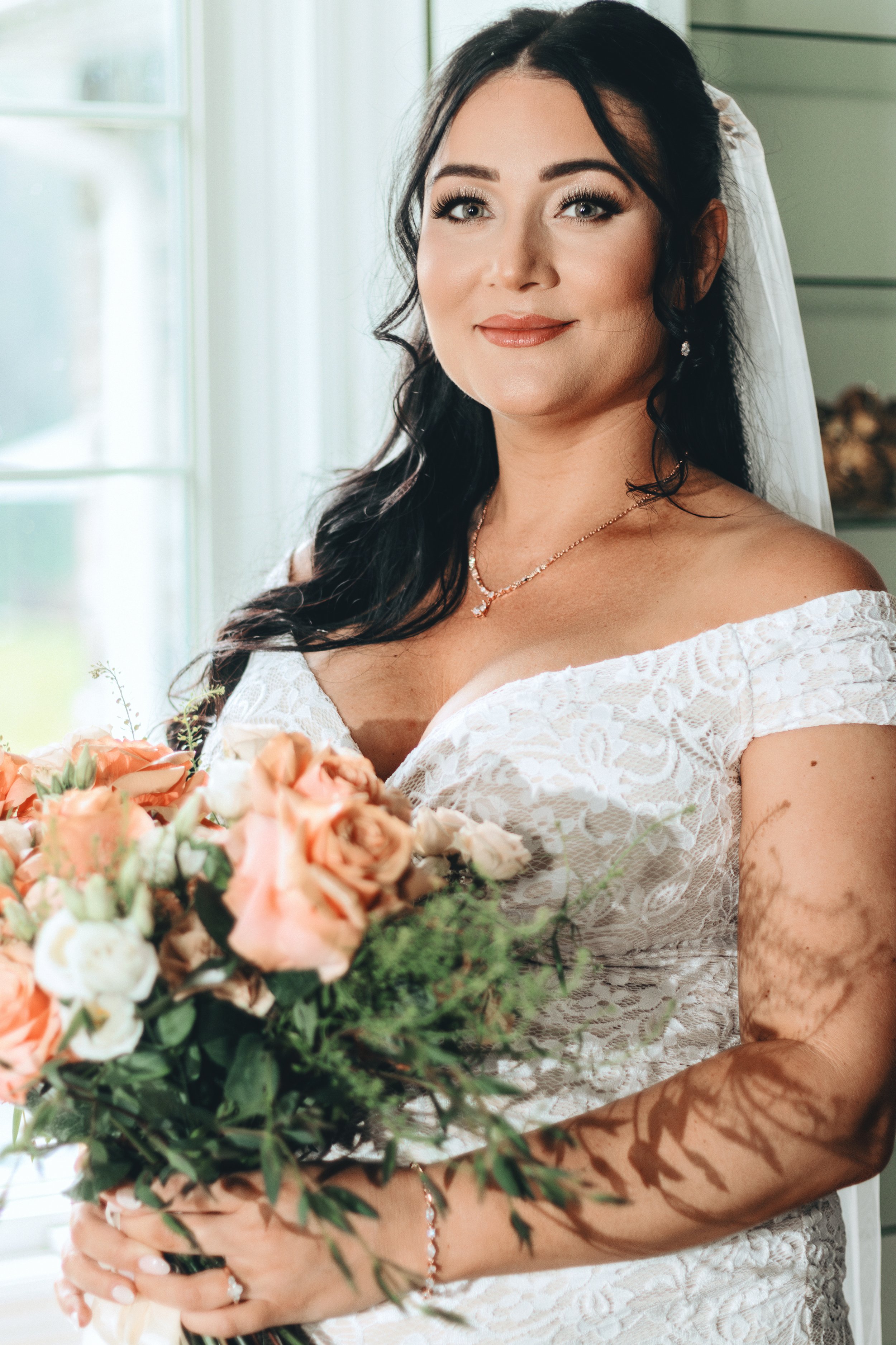 A bride with dark hair and light skin, wearing a white lace off-shoulder wedding dress, holding a bouquet of pink and white roses, standing indoors near a window with natural light.