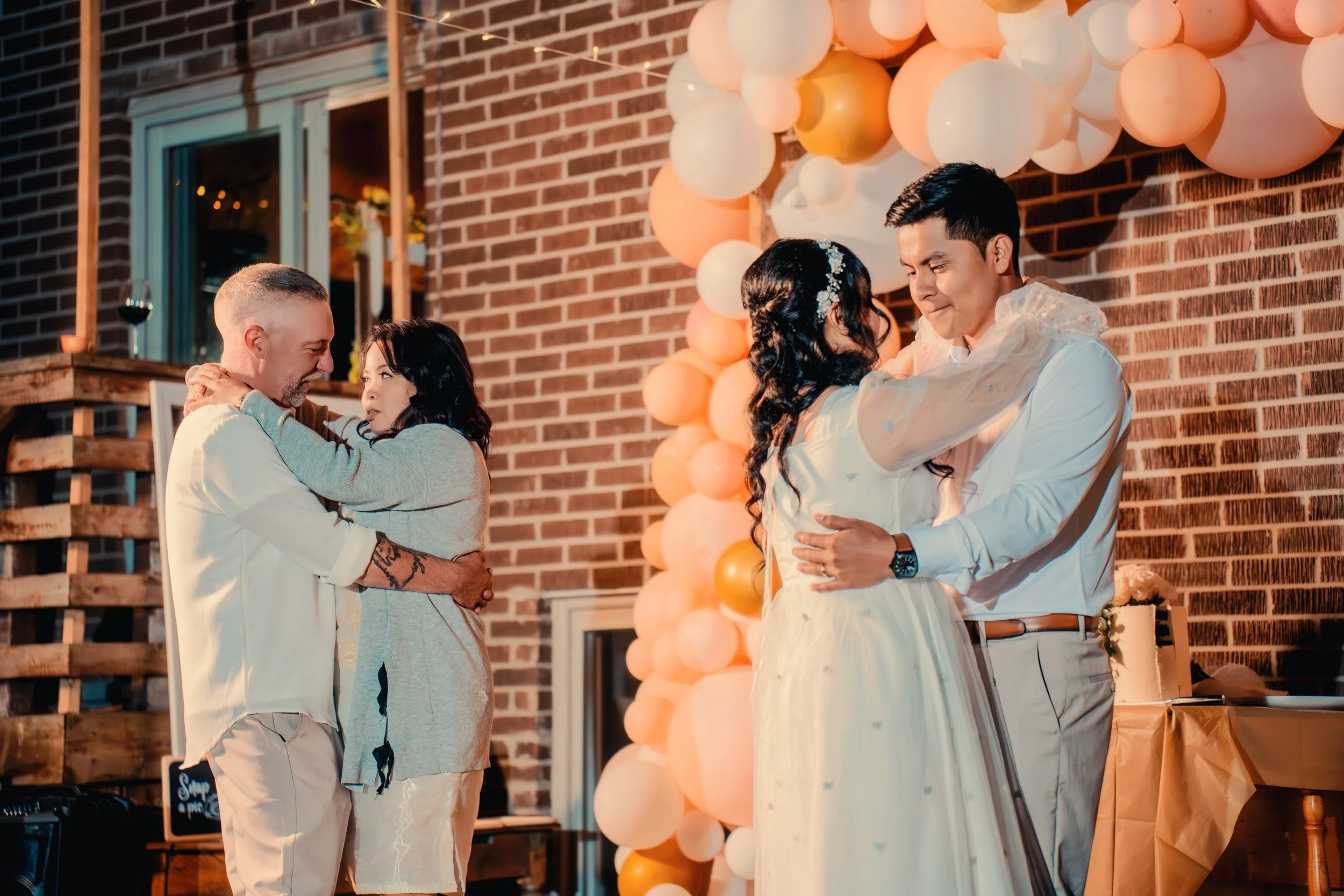 Two couples dancing closely at a wedding reception with a brick wall and balloon decoration in the background.