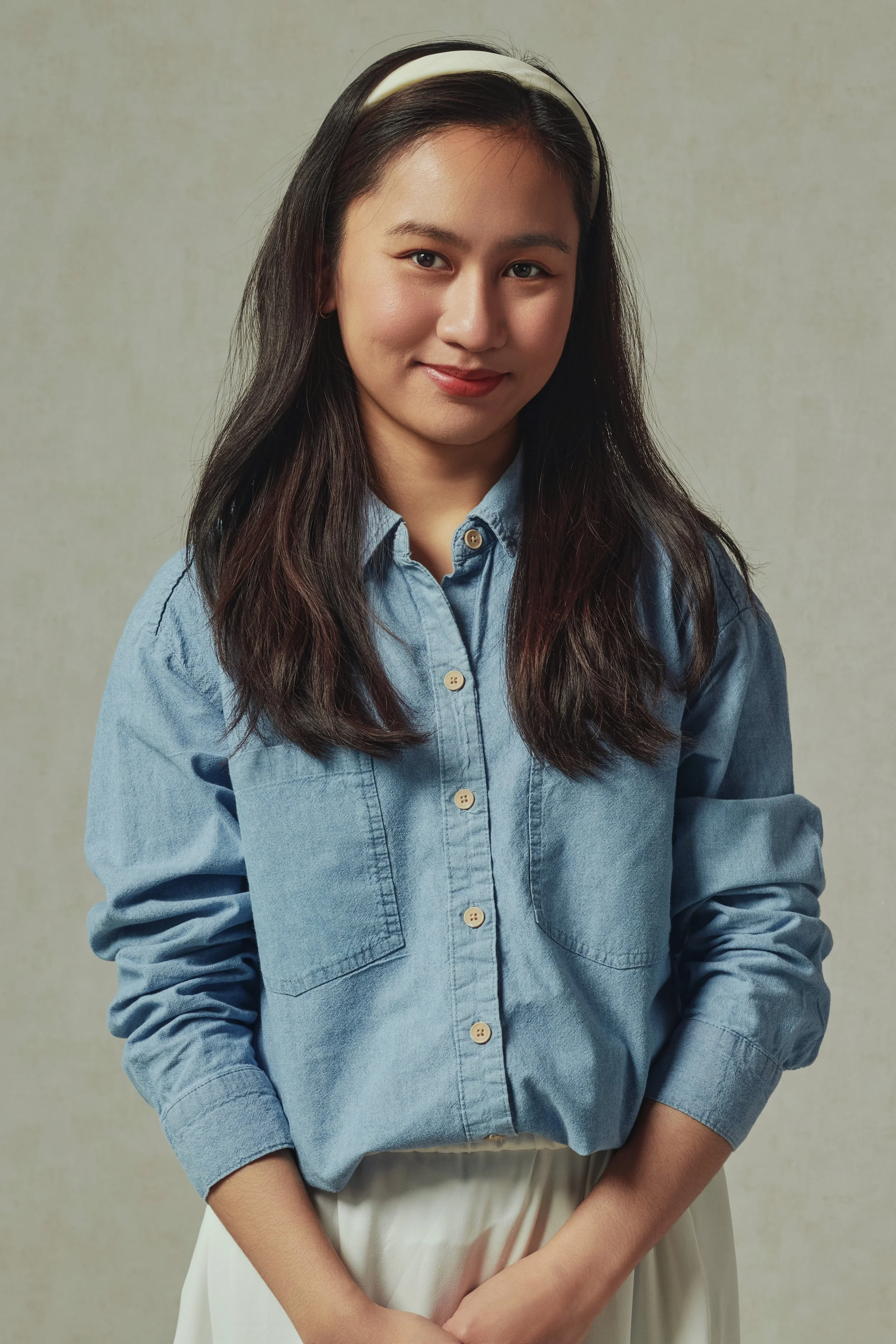 A young woman with long dark hair wearing a white headband, a blue denim button-up shirt, and cream-colored pants standing against a neutral background.