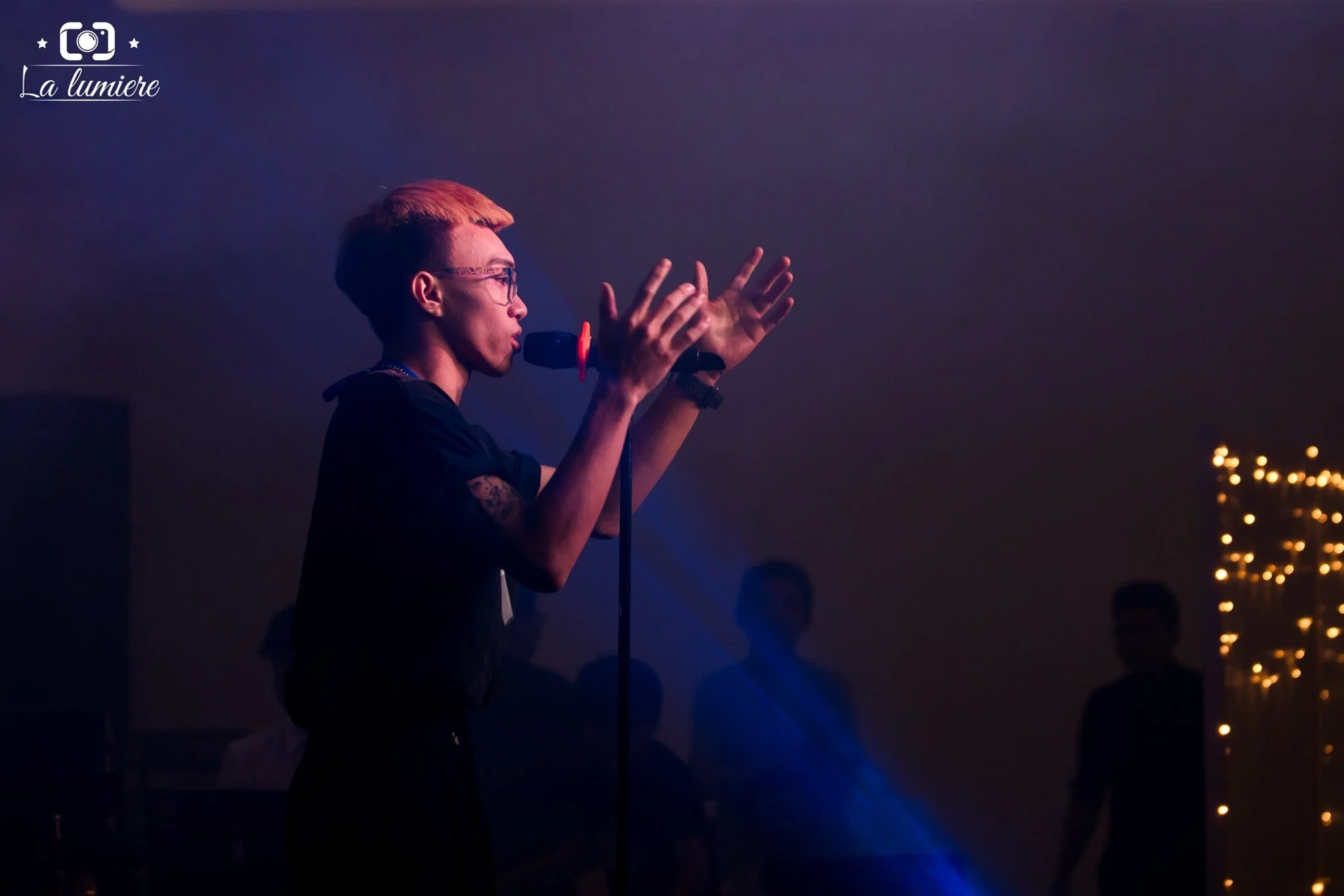 A young person with orange hair and glasses singing into a microphone on stage, with a dark background and a few blurred audience members visible.