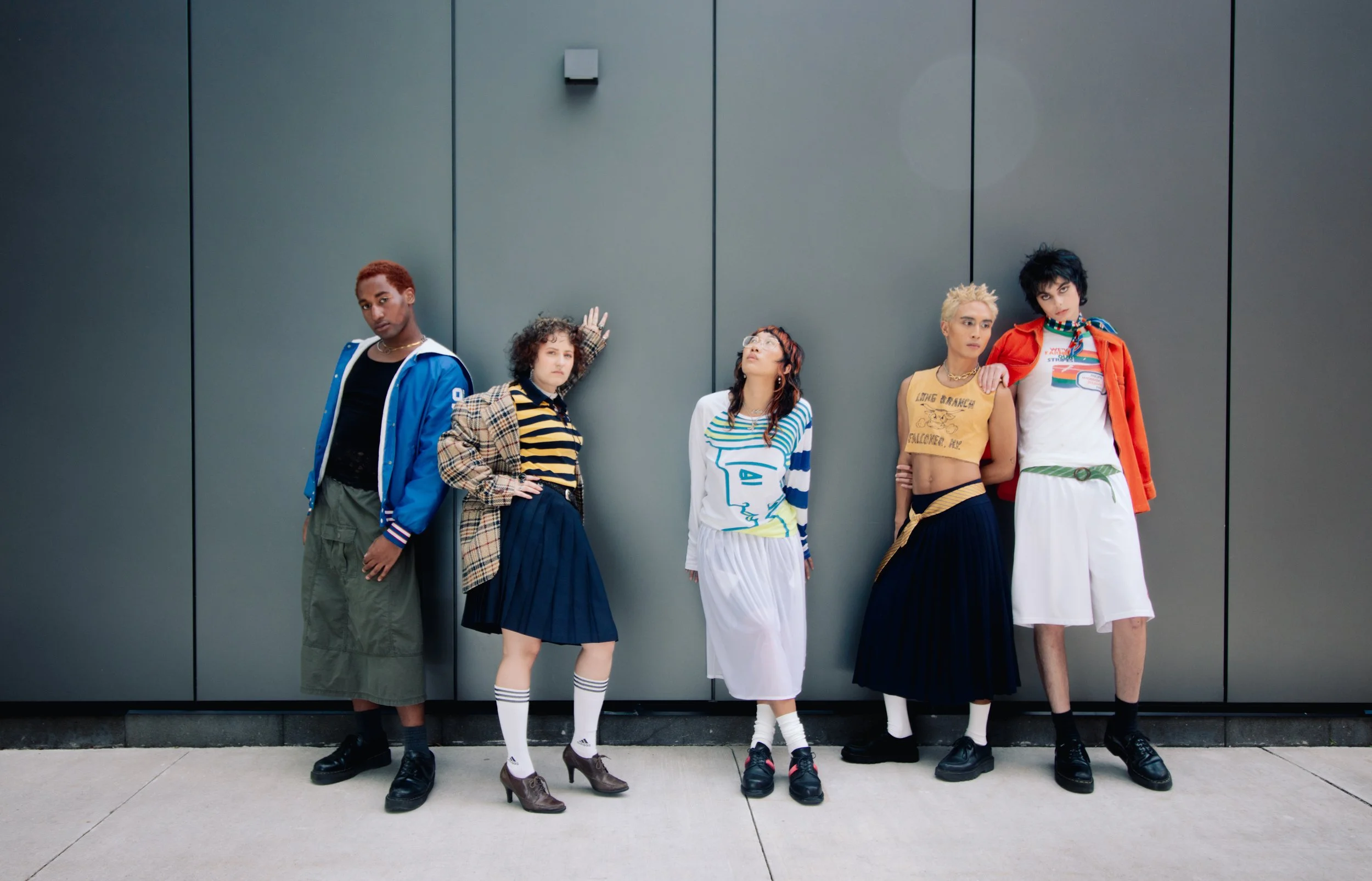 Five diverse young people standing against a gray wall, dressed in colorful, trendy fashion styles.