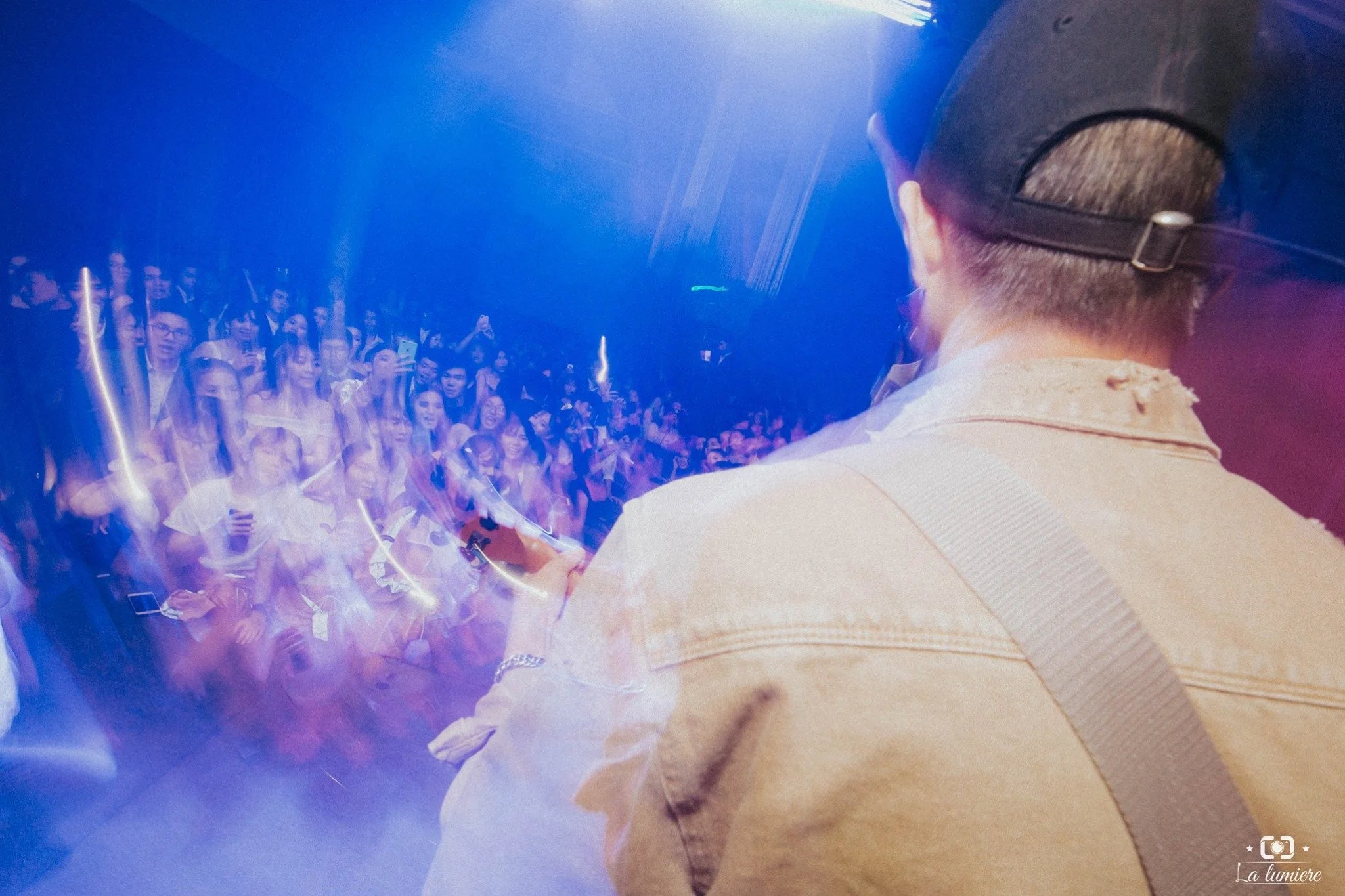 A person wearing a backward cap and beige jacket is on stage performing or speaking to a crowd. The audience appears excited, with many holding up phones, and the scene is illuminated with colorful stage lights.