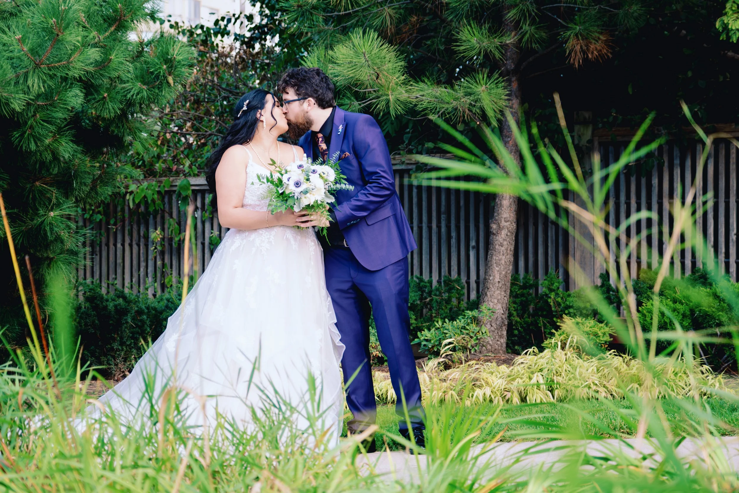 A bride and groom sharing a kiss outdoors, surrounded by greenery and trees, with the bride holding a bouquet of white and purple flowers.