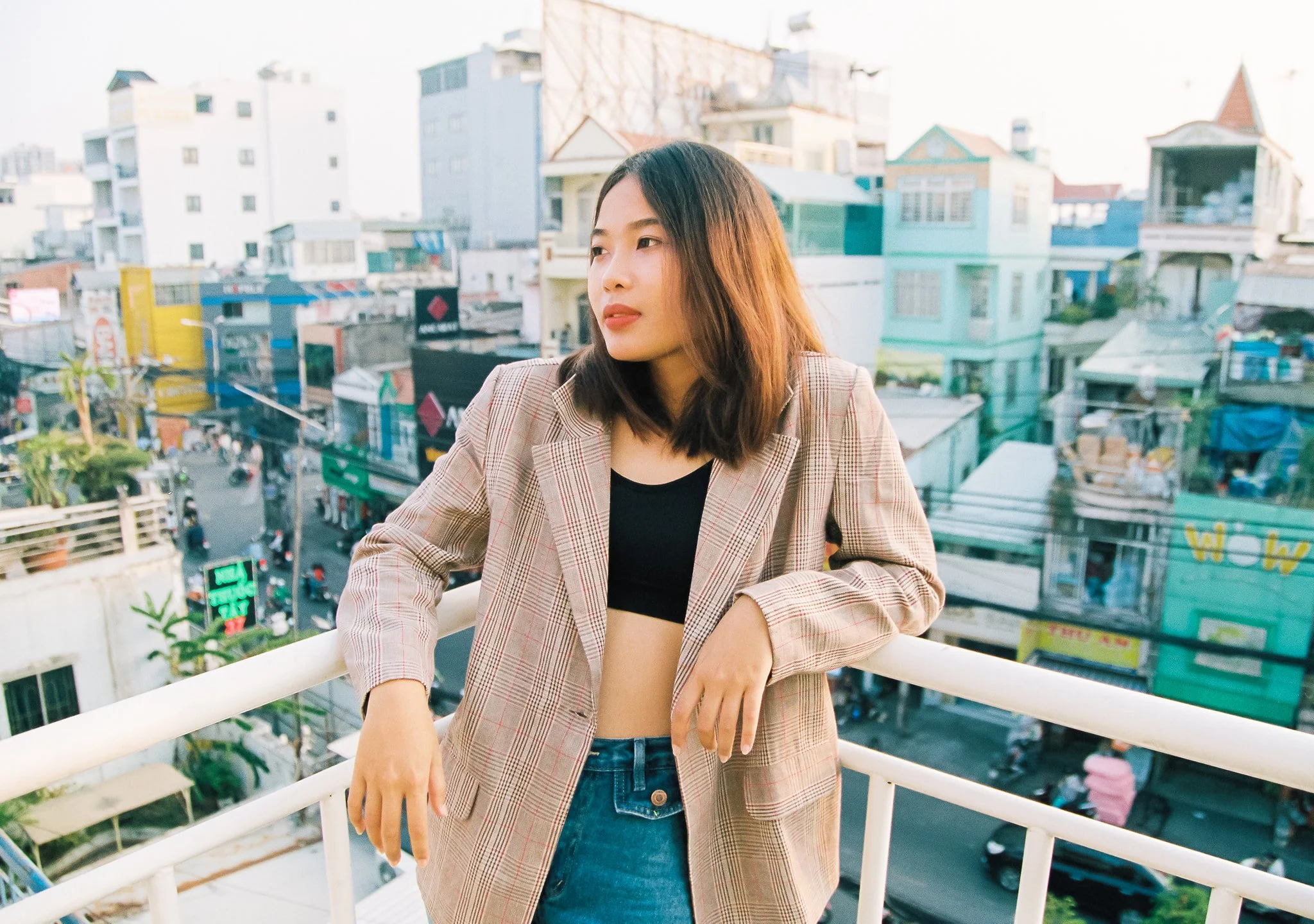 A young woman with shoulder-length hair wearing a plaid blazer over a black crop top and jeans, leaning on a white railing on a rooftop in an urban area with buildings and street below.