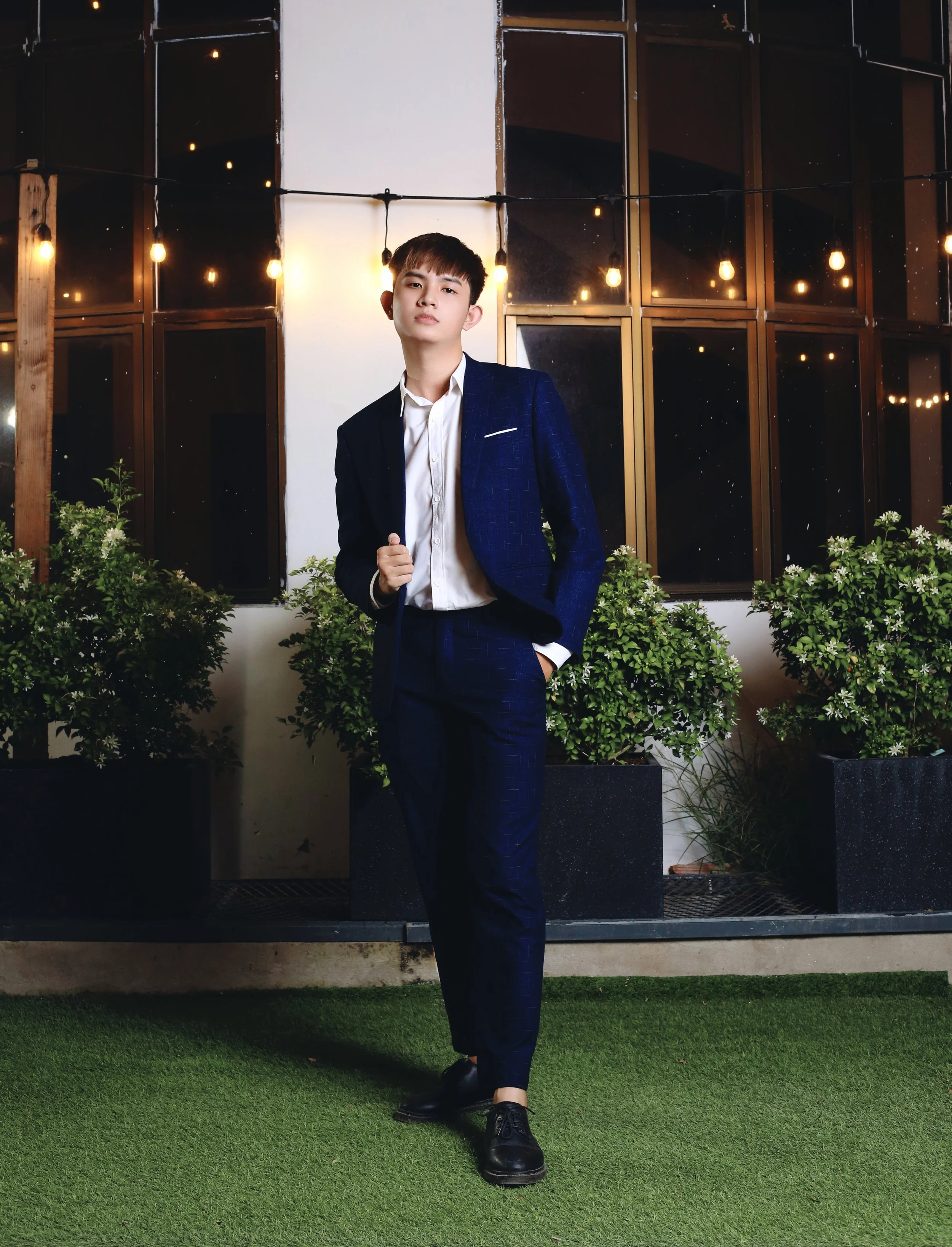 Young man in a navy suit with a white shirt, standing outdoors at night with string lights, potted plants, and windows in the background.