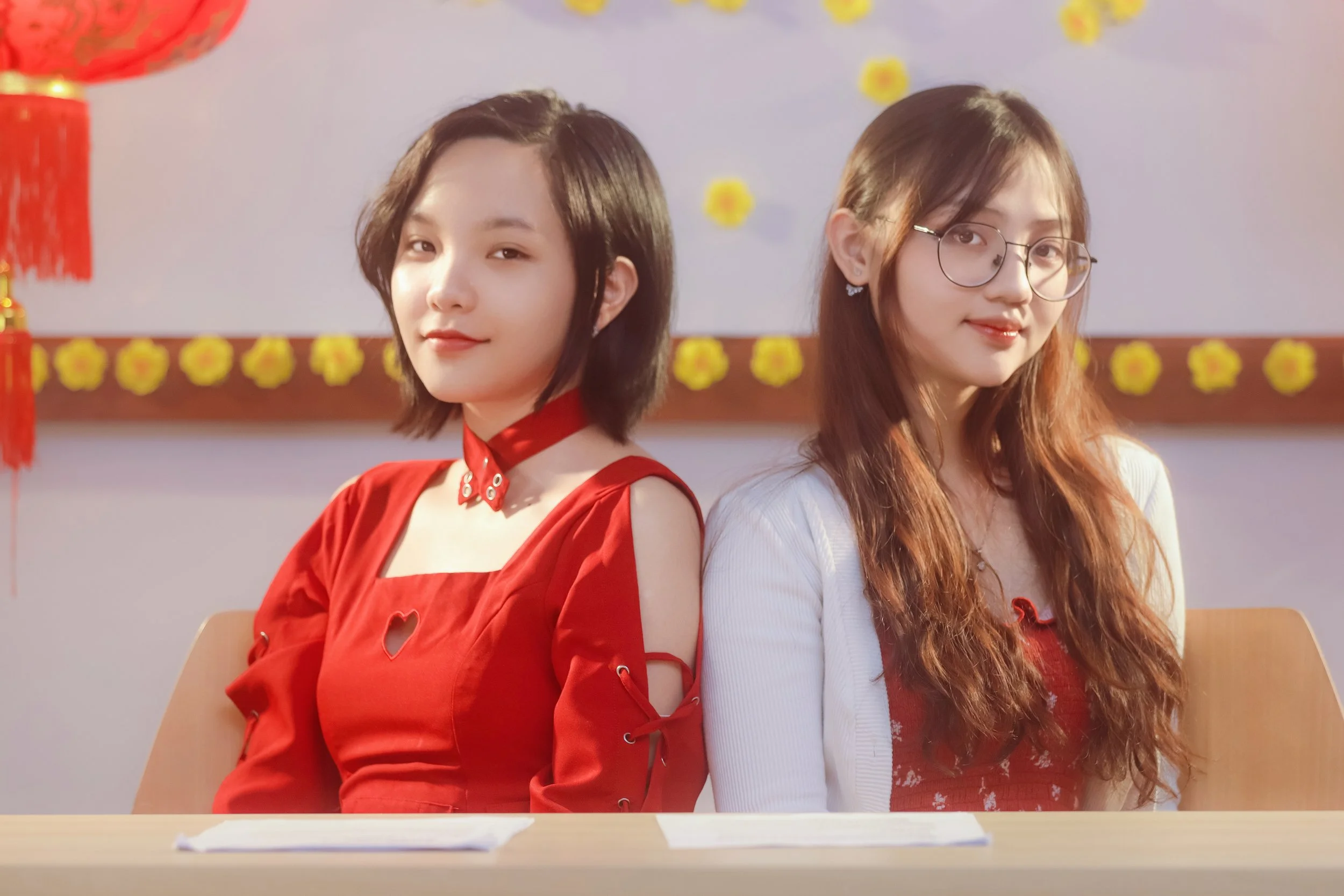 Two young women sitting back to back at a table during a festive celebration, one wearing a red dress with cut-out shoulders and a choker, the other wearing glasses and a white cardigan over a red dress, with decorative yellow flowers and paper lante