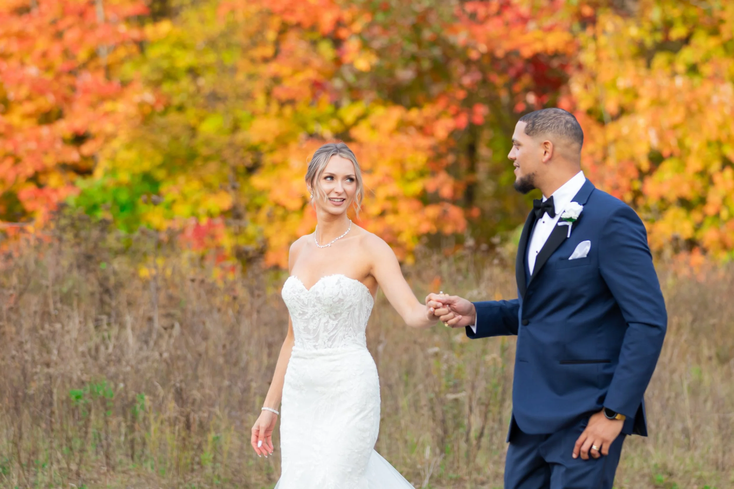 A bride and groom holding hands outdoors during fall, with colorful autumn trees in the background.