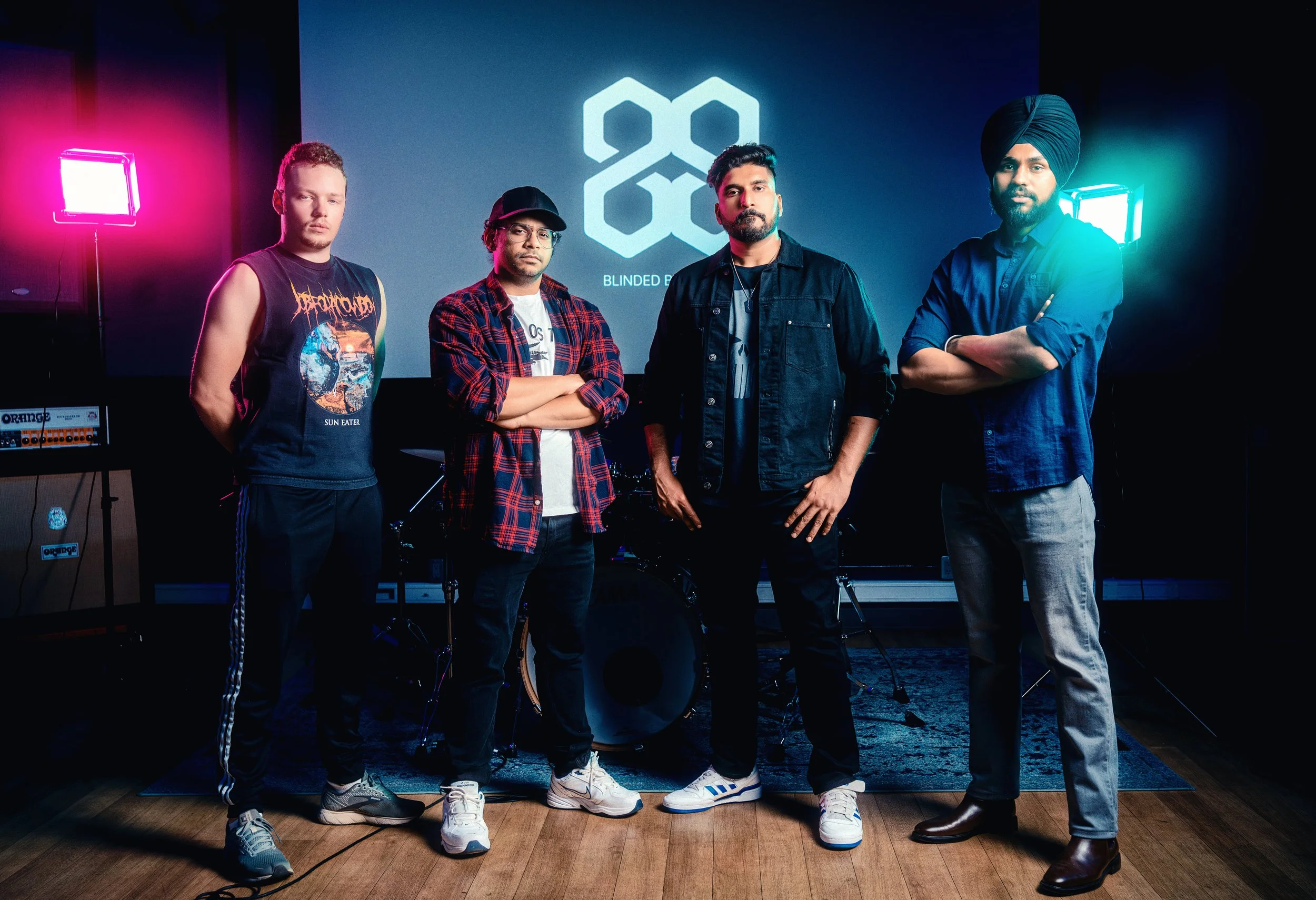 Four men standing in front of a screen with a logo that reads 'BLINDED RABBIT'. The men are dressed casually, with one wearing a sleeveless band t-shirt, another in a baseball cap and plaid shirt, the third in a denim jacket, and the fourth in a turb
