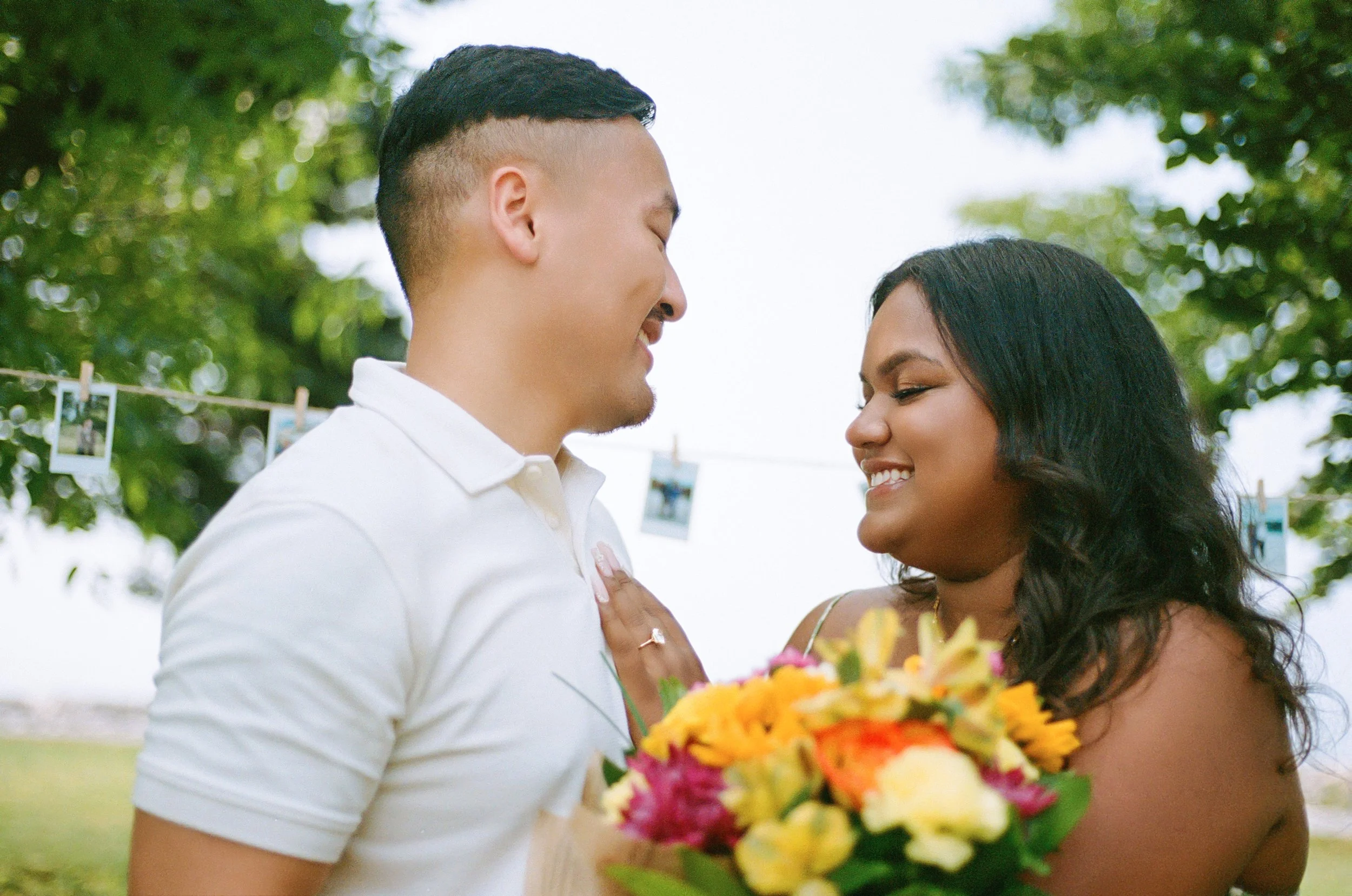 A joyful couple stand close together outdoors, smiling at each other, with a bouquet of colorful flowers. The woman has dark hair and tan skin, and the man has short dark hair and light skin. Green trees and a string of photos in the background sugge