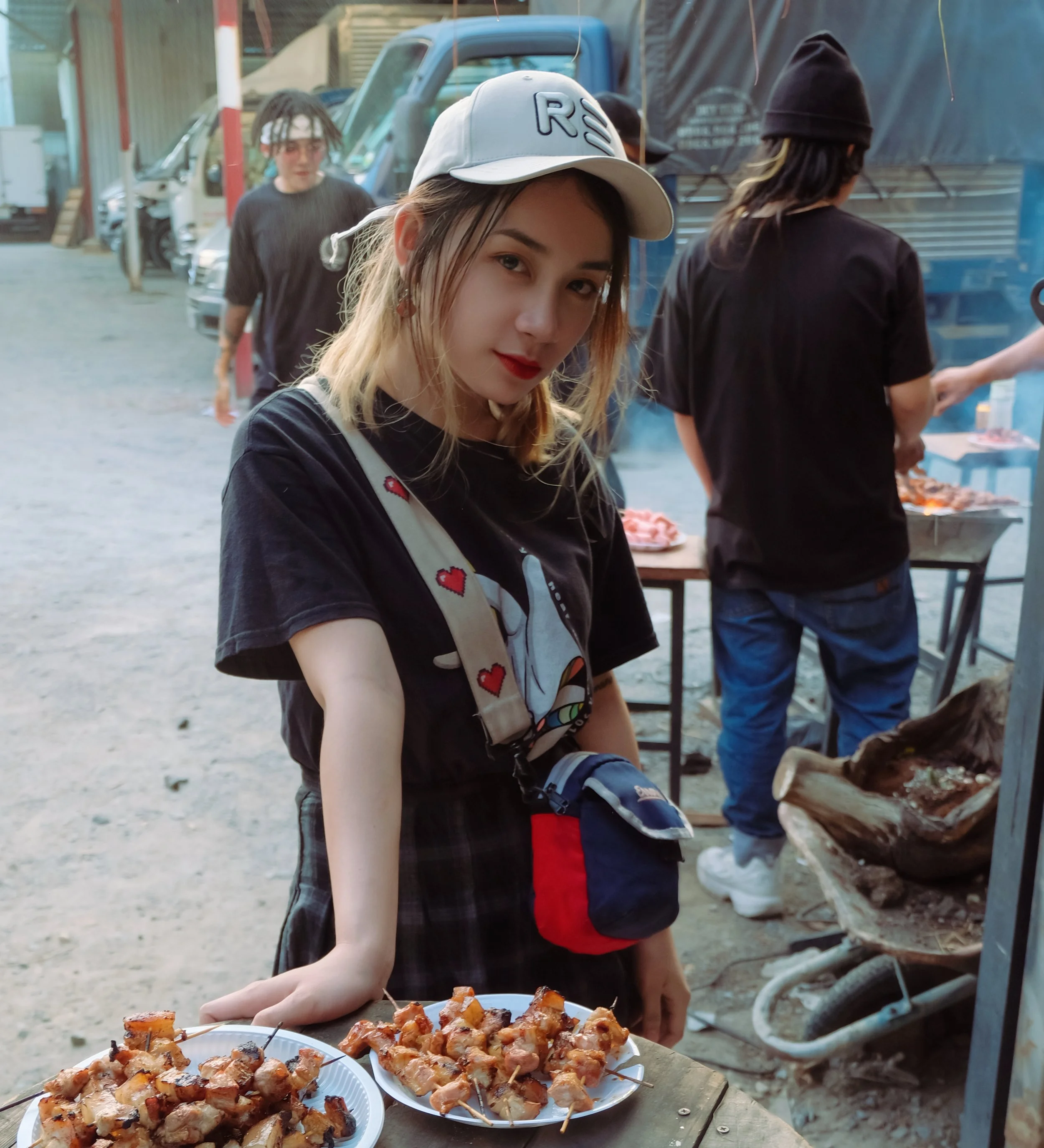 A young woman with blonde hair, wearing a white cap and black t-shirt, stands outdoors at a food stall with plates of skewered grilled meat. There are other people and vehicles in the background.