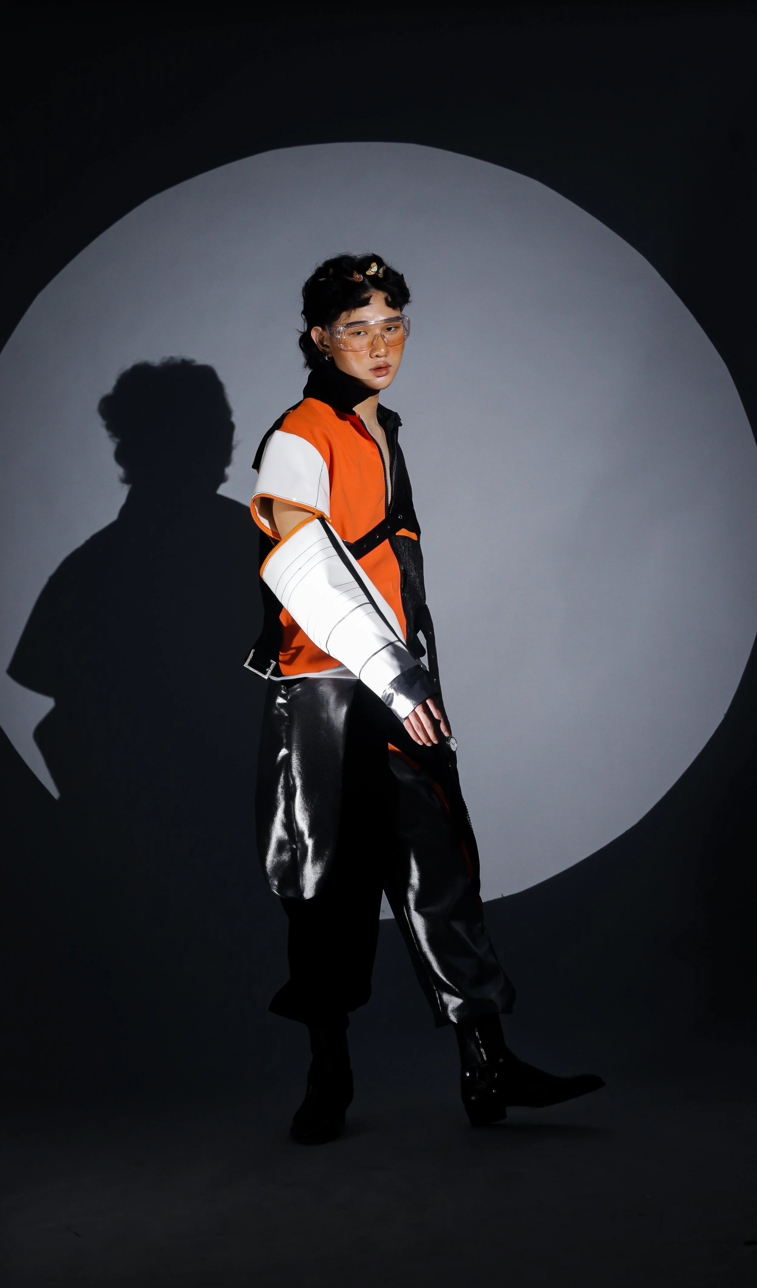 A person in futuristic fashion standing in front of a gray background with a circular spotlight, wearing a multicolored jacket, shiny black pants, and protective glasses.