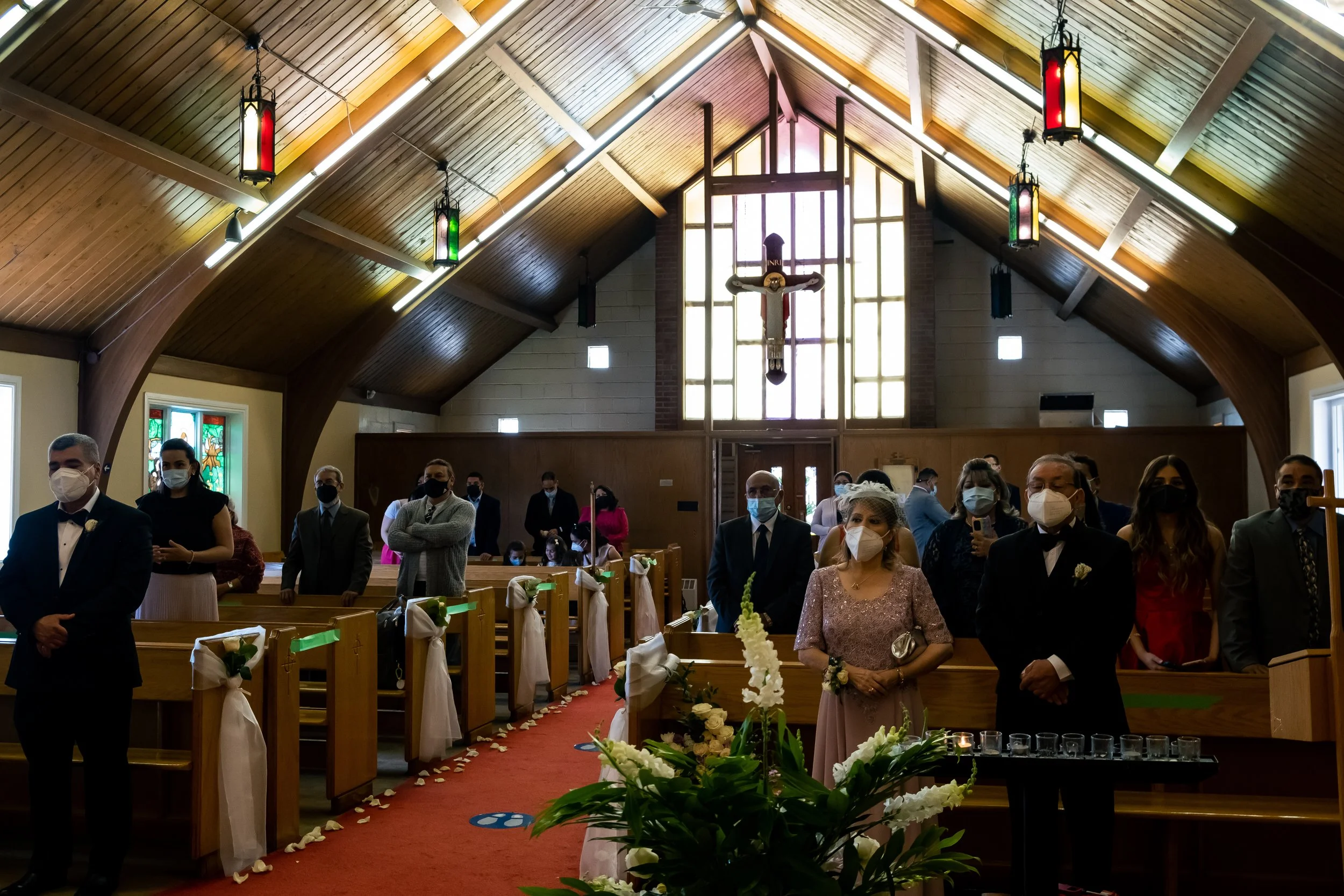Couples and guests attending a wedding ceremony inside a church with wooden ceilings, stained glass windows, and a large cross at the altar, all wearing face masks.