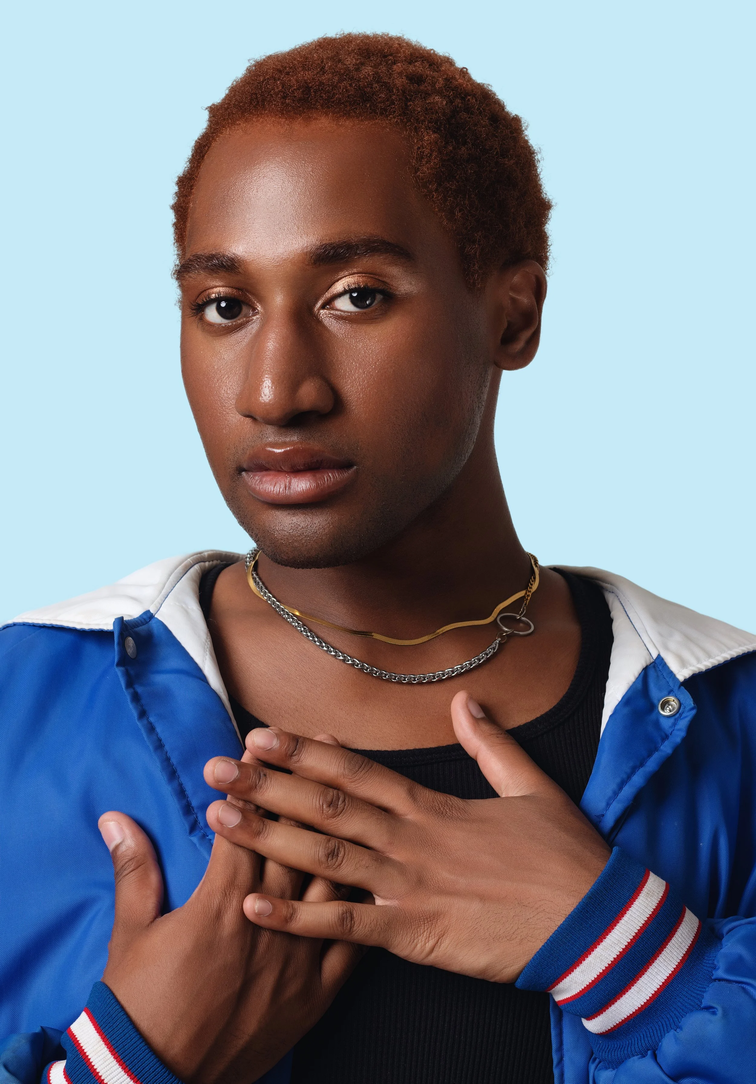 A person with short, reddish-brown hair and brown skin gazes at the camera, wearing layered necklaces and a blue and white jacket over a black shirt, with their hands over their chest against a light blue background.