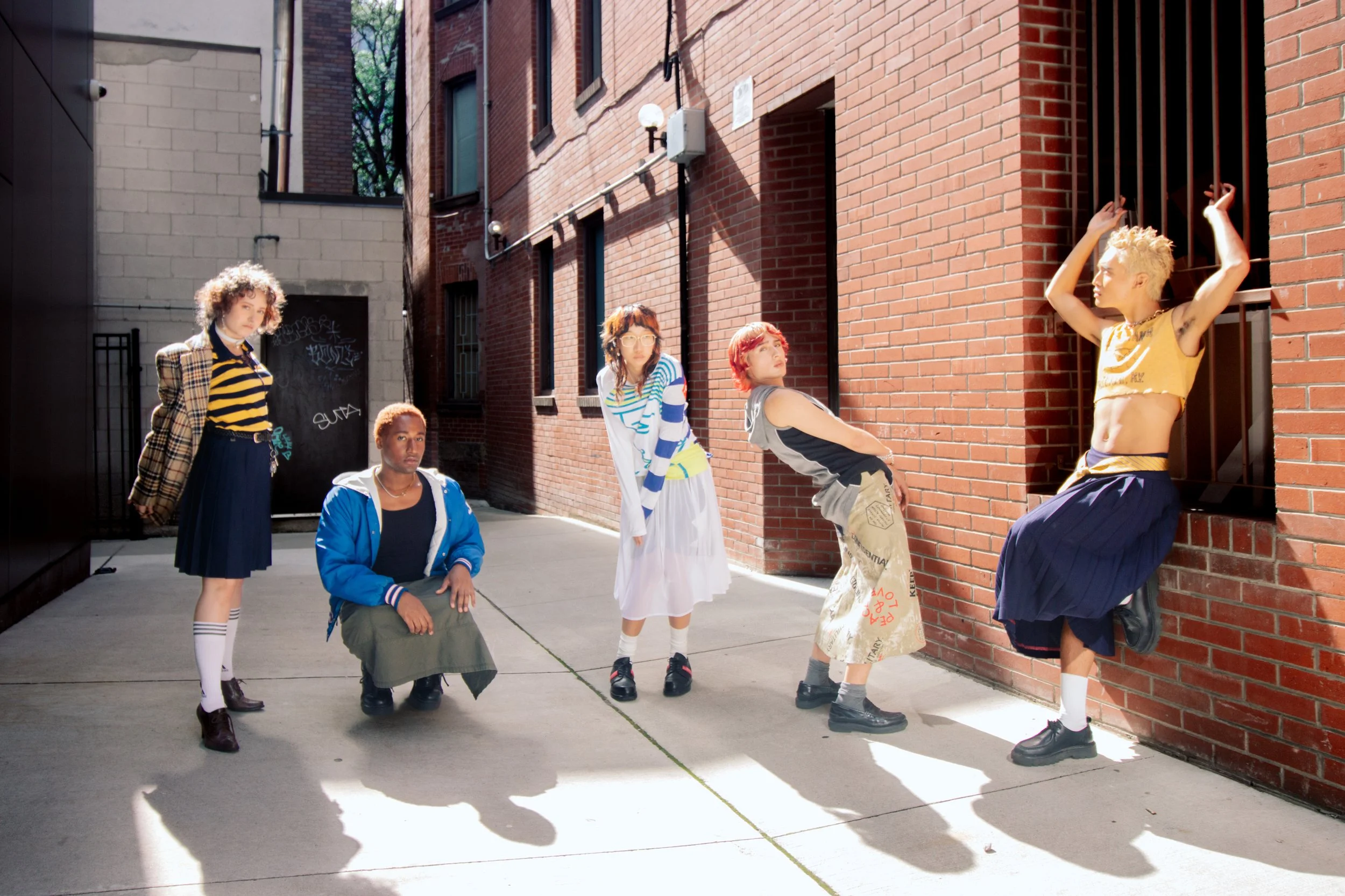 Five young women posing in an alleyway with brick buildings. One is sitting, two are standing, and two are leaning against a brick wall, dressed in casual and trendy clothing.
