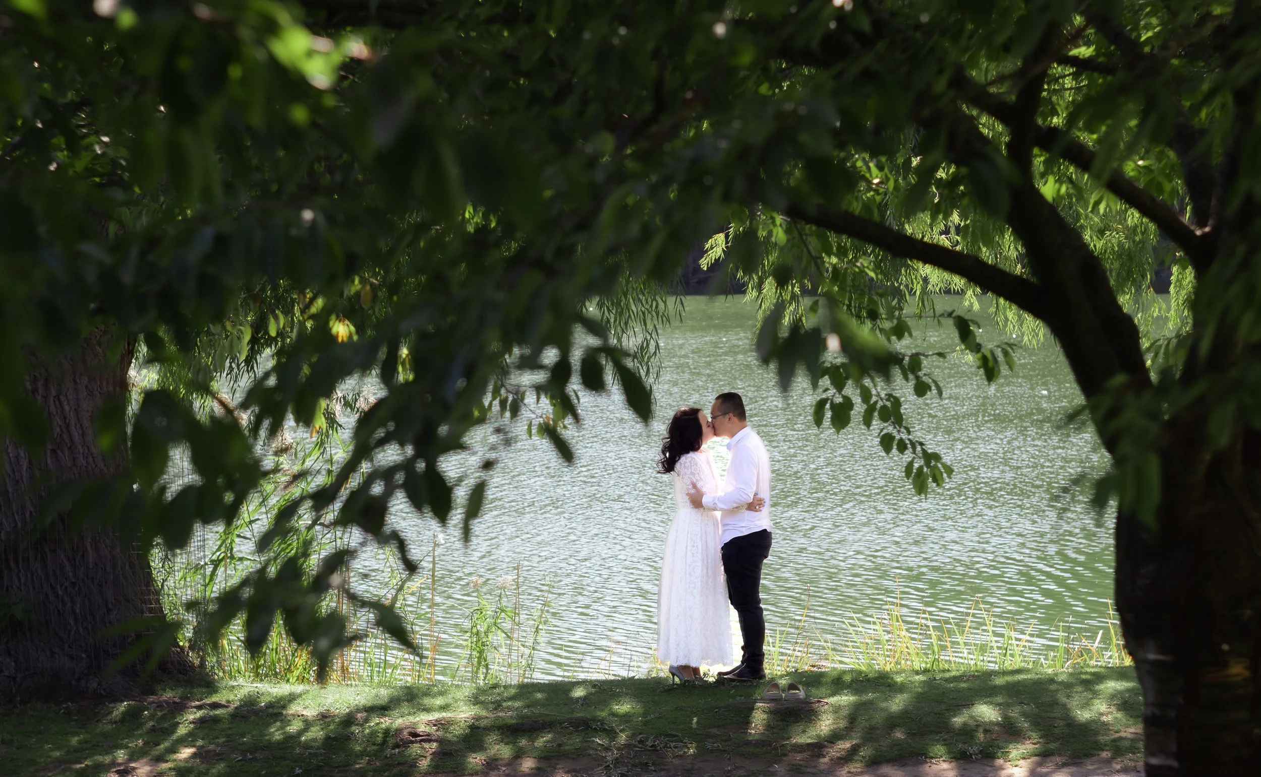 A couple dressed in white, standing by a lake under trees, about to kiss.